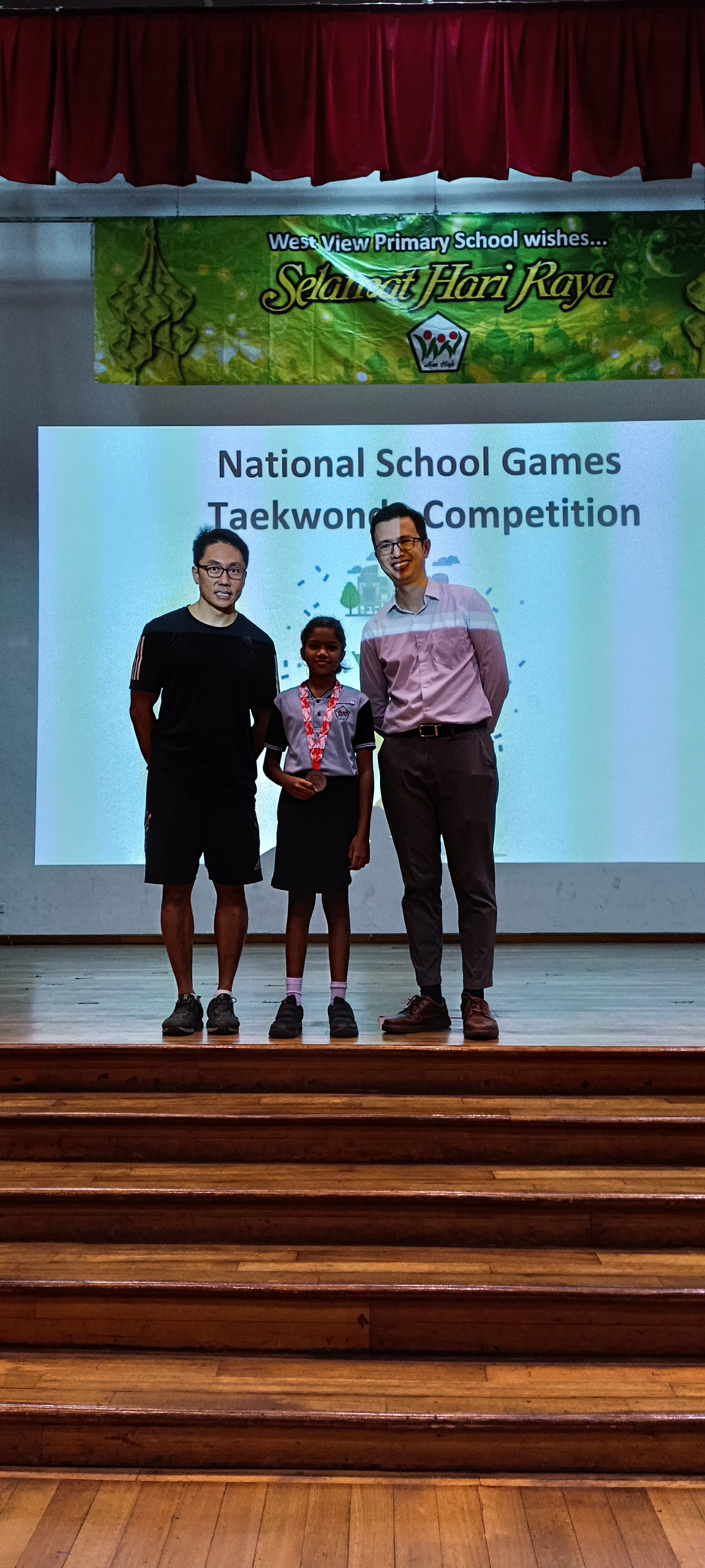 Two adults and a child with a medal stand on a school stage in front of a National School Games Taekwondo Competition screen.