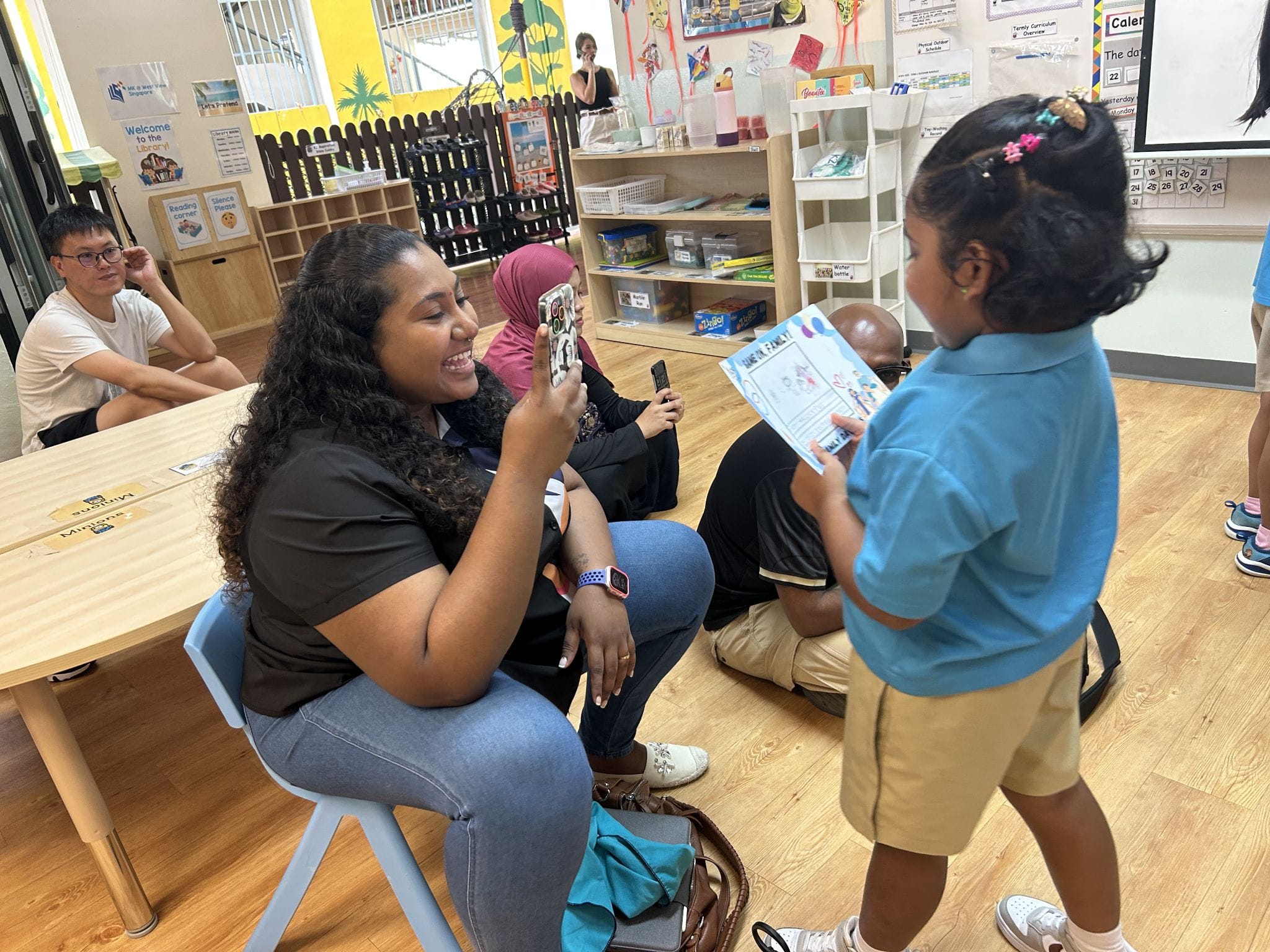 A woman smiles and photographs a young girl holding a drawing in a colorful classroom with adults seated nearby.