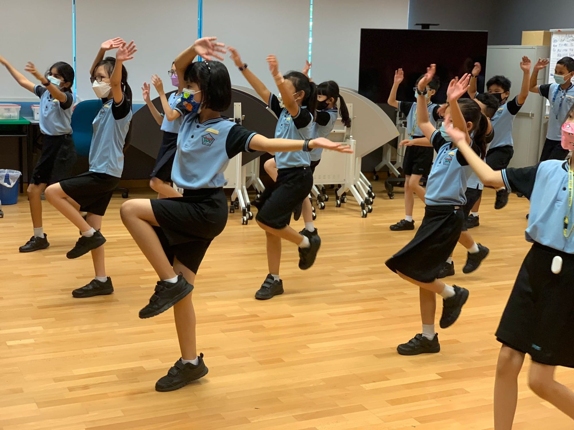 Students in blue uniforms and masks perform a synchronized dance with raised arms and lifted knees on a wooden floor.