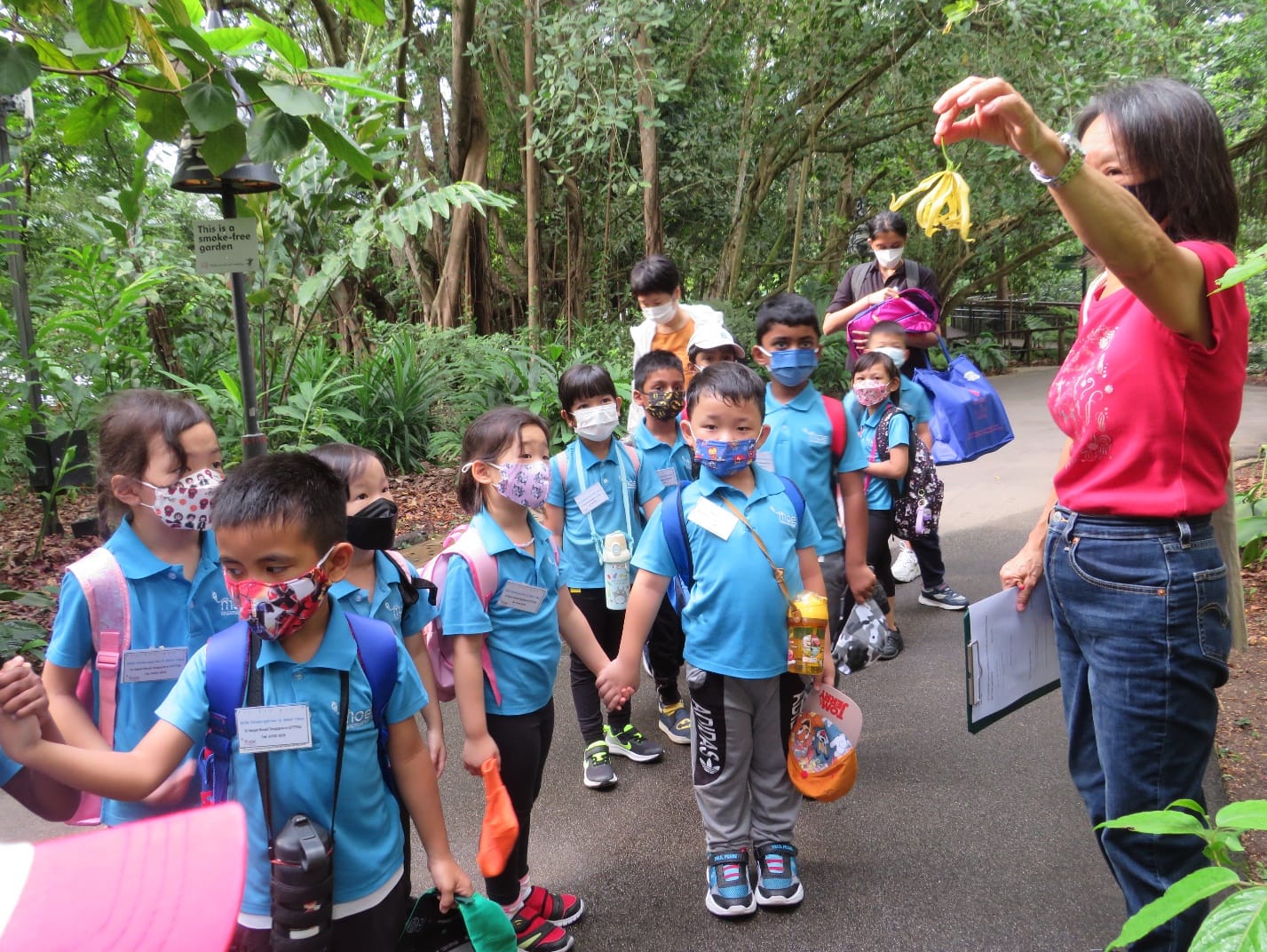 A woman in red holds up a yellow flower for masked children in blue uniforms on a garden path.