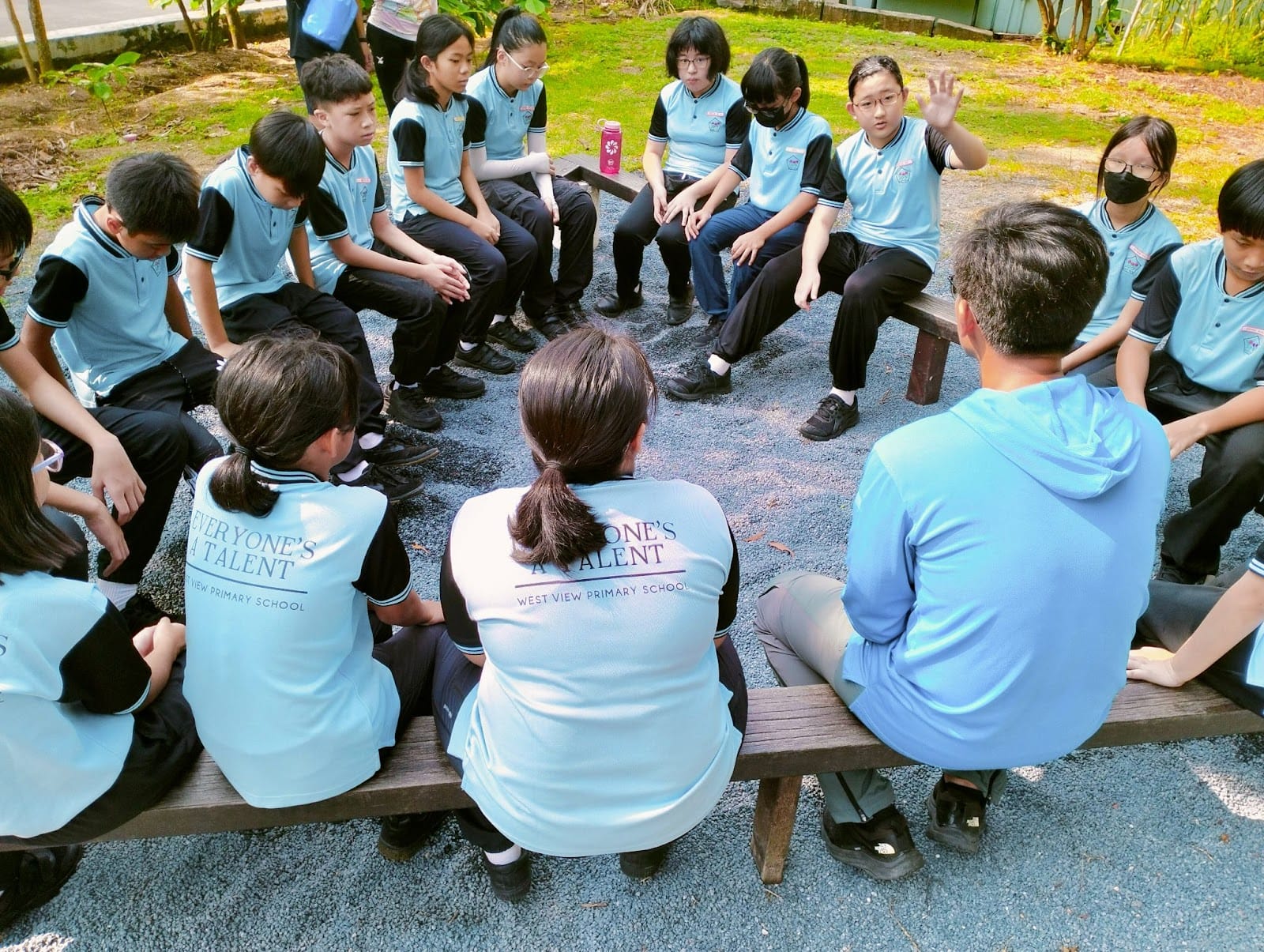 Students in light blue West View Primary School uniforms sit in a circle on outdoor benches having a group discussion.