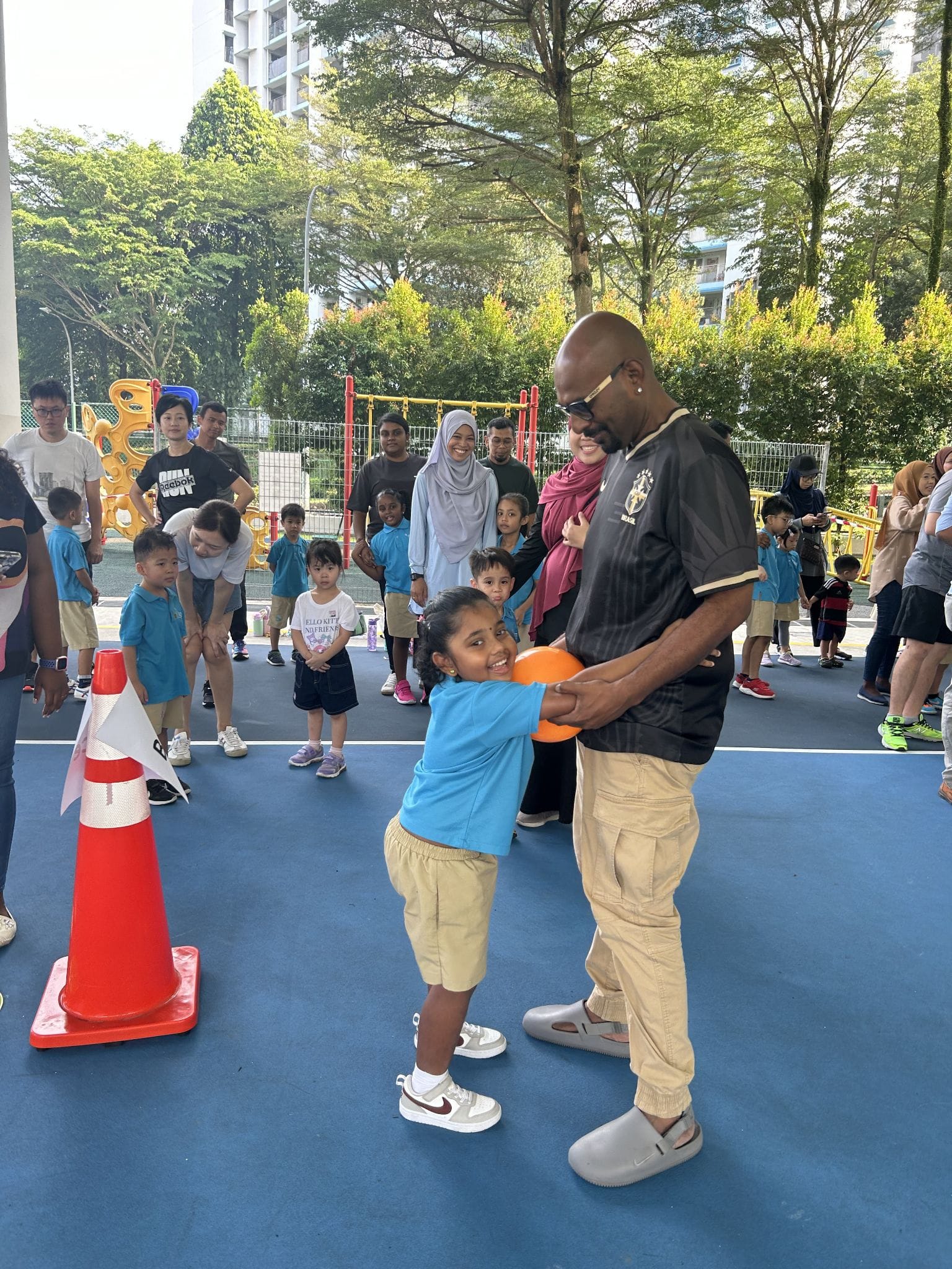 A young girl in a blue shirt hugs a man holding an orange balloon at an outdoor school sports event with children watching.