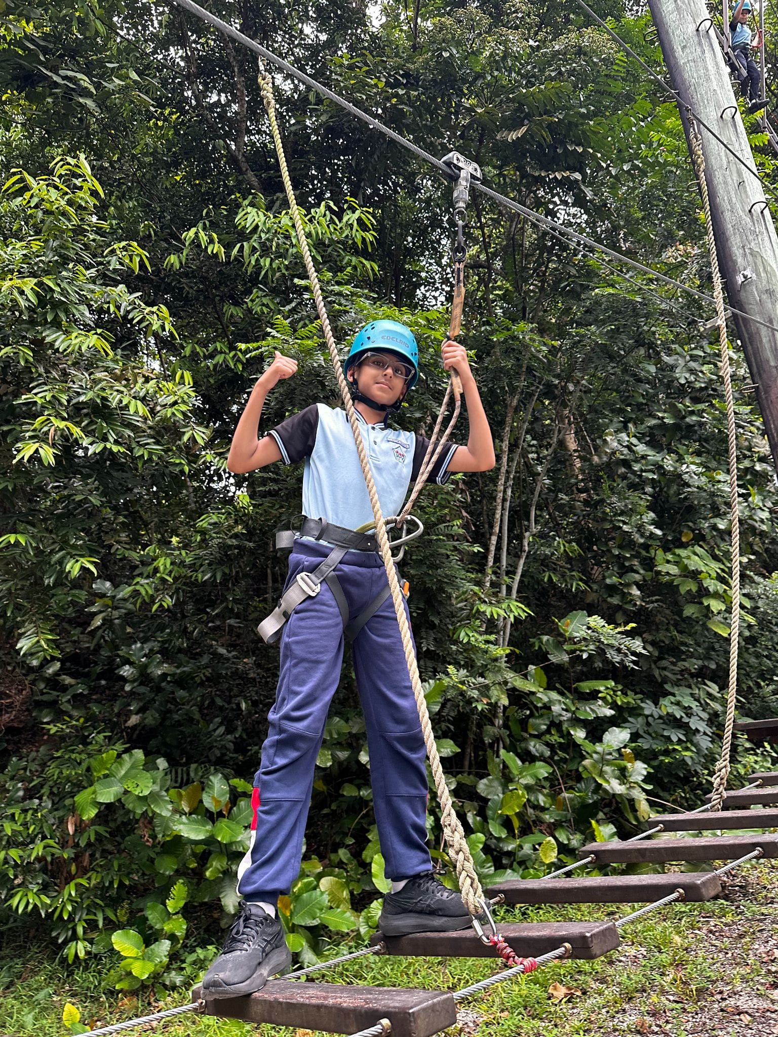 A child wearing a blue helmet and harness stands on a rope ladder course surrounded by green trees.