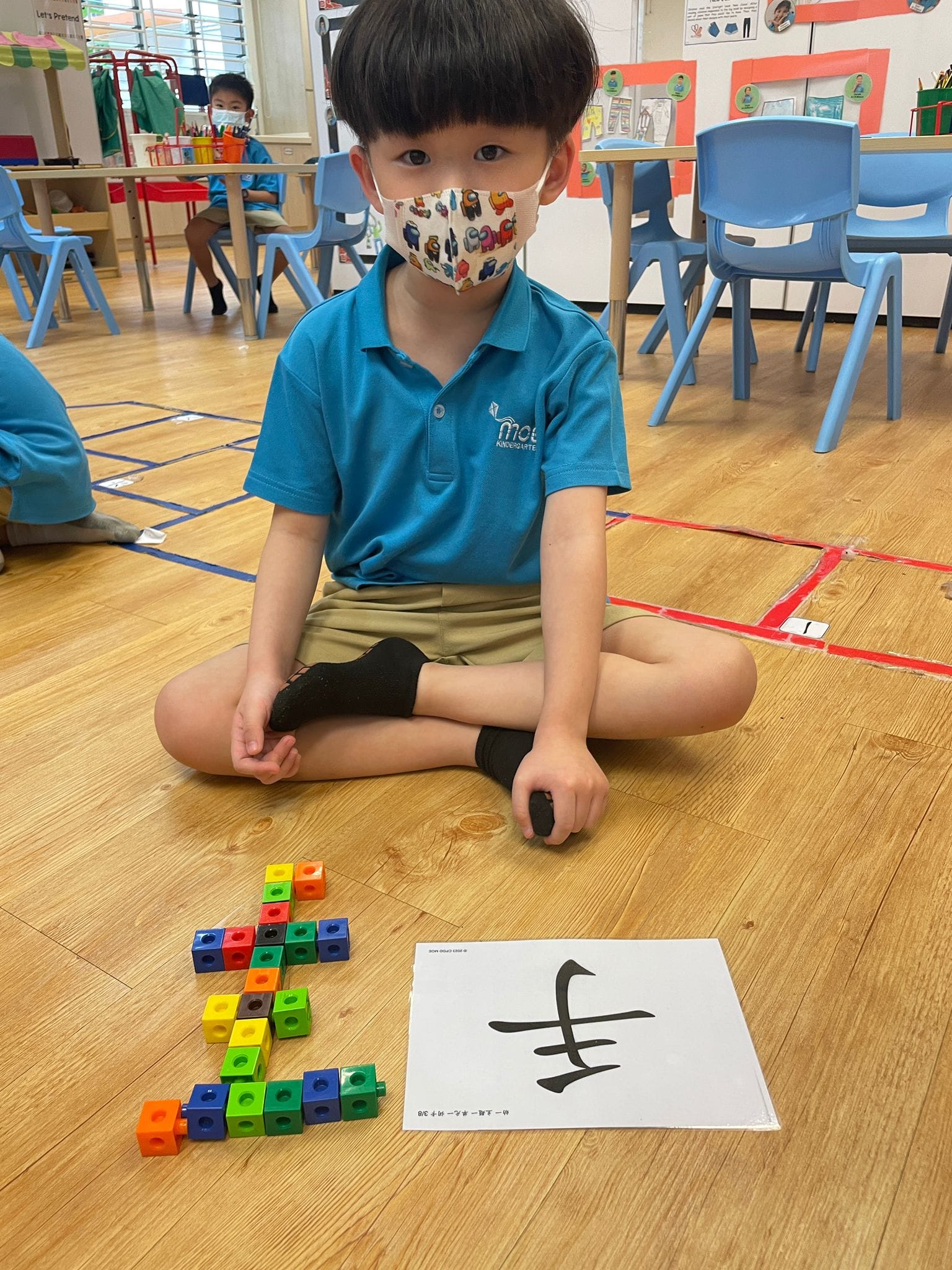 Young masked child sits cross-legged on classroom floor beside colorful snap cubes arranged as a Chinese character shown on a card.