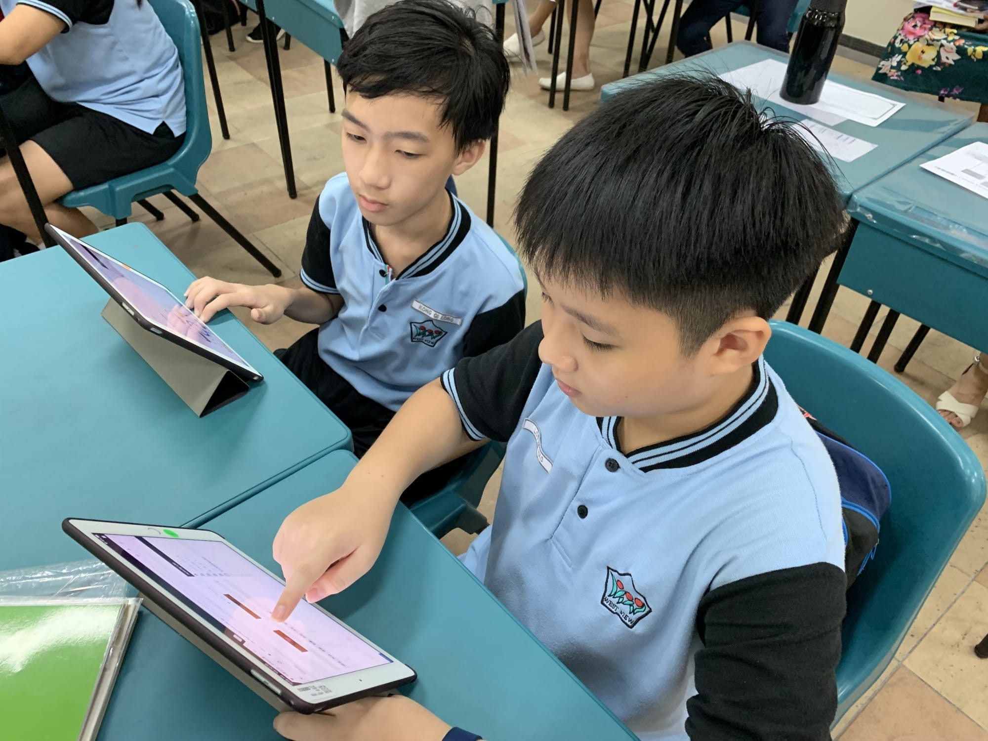 Two boys in blue school uniforms sit at teal desks, each using a tablet in a classroom.