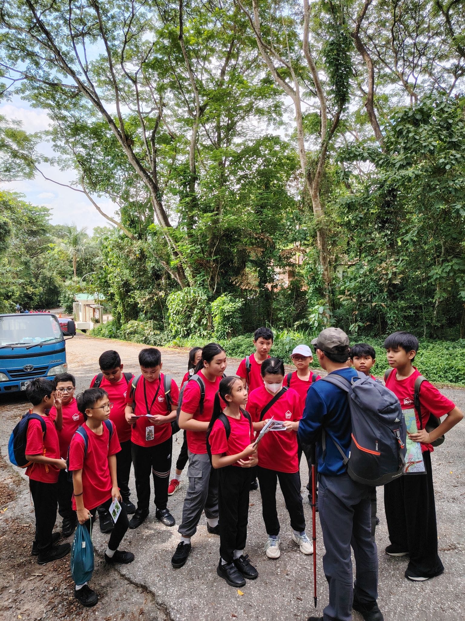 A group of children in red shirts with backpacks gather around an adult guide holding a map outdoors near trees.