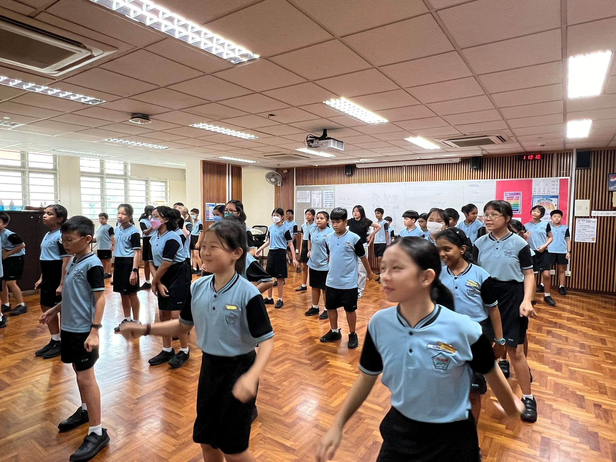 A large group of children in light blue and black uniforms move around on a wooden floor in a classroom.