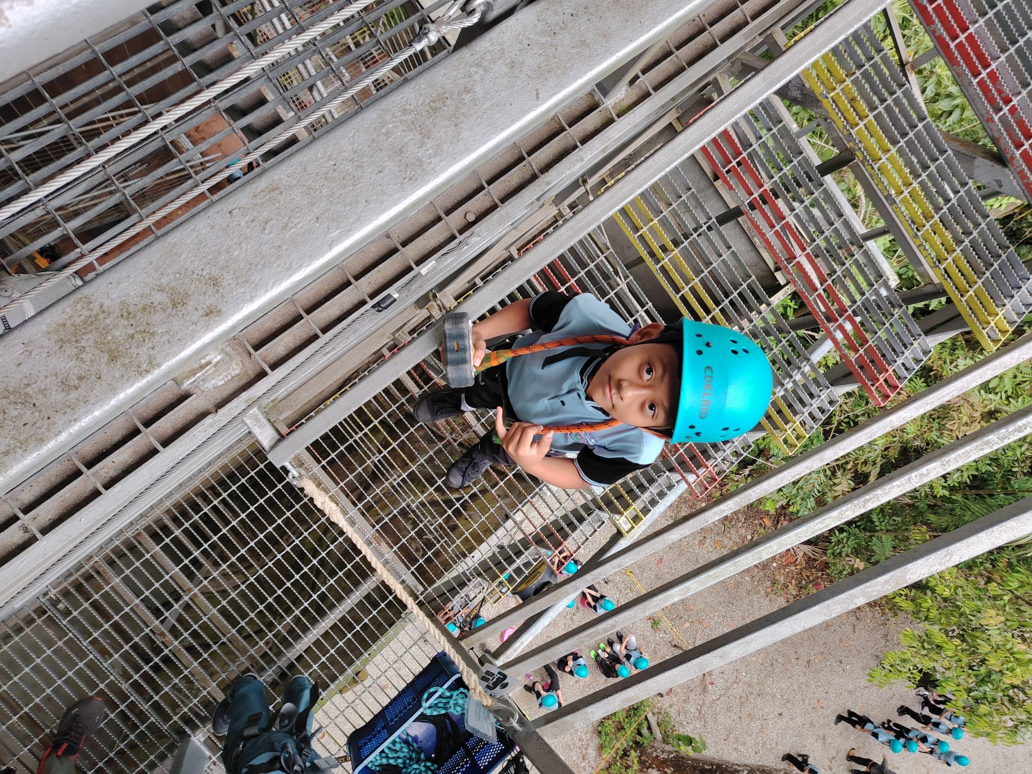 A child wearing a blue helmet and harness stands on a metal grate platform at a high ropes or zip-line course.