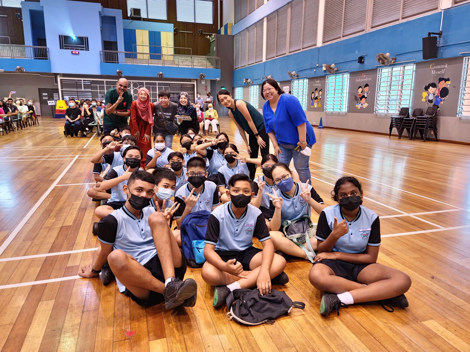 A group of masked students in light blue uniforms sit on a school hall floor, posing with adults behind them.