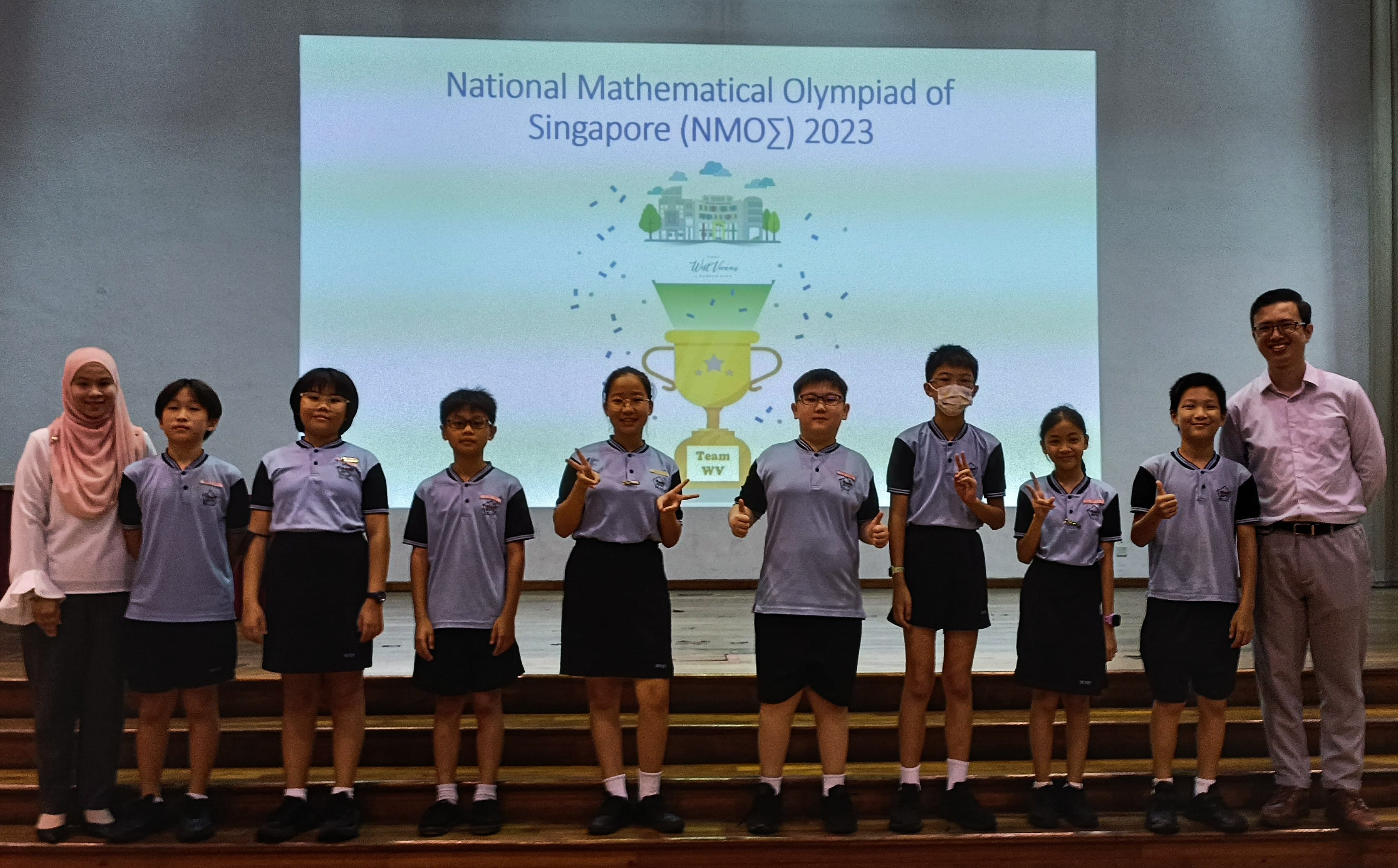 Students and teachers pose in front of a National Mathematical Olympiad of Singapore 2023 presentation slide.