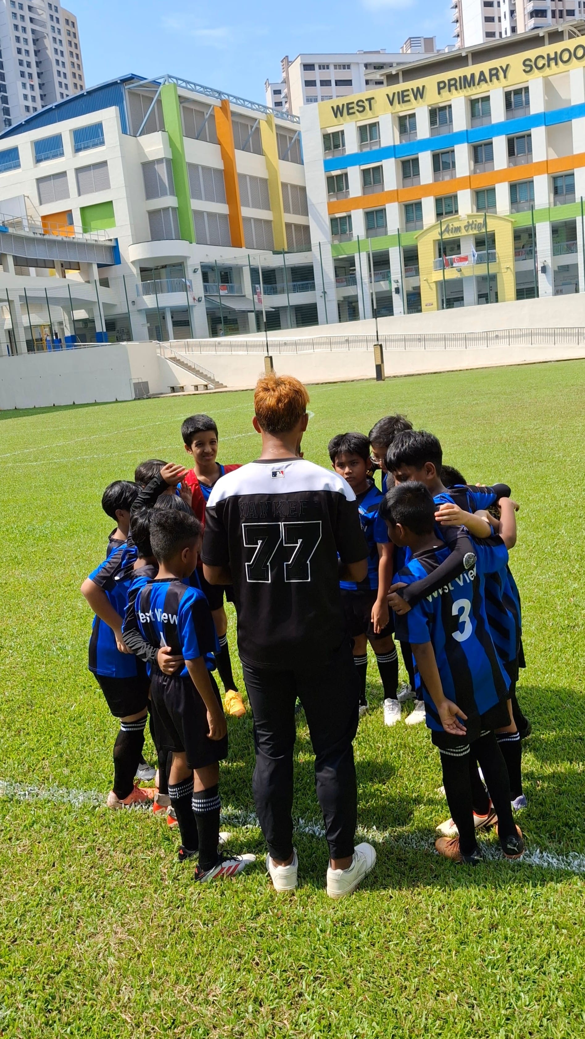Youth soccer team huddles on grass field near a building labeled West View Primary School.