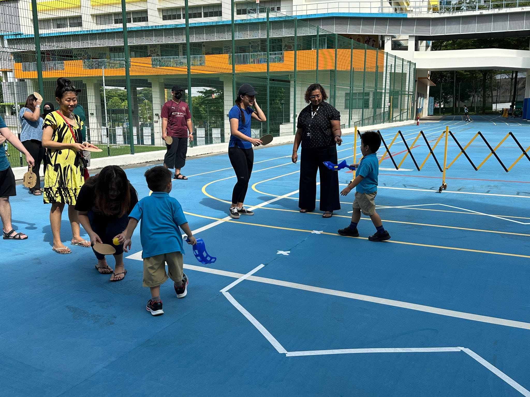 Adults and young children play with paddles and balls on a blue outdoor court at a school.
