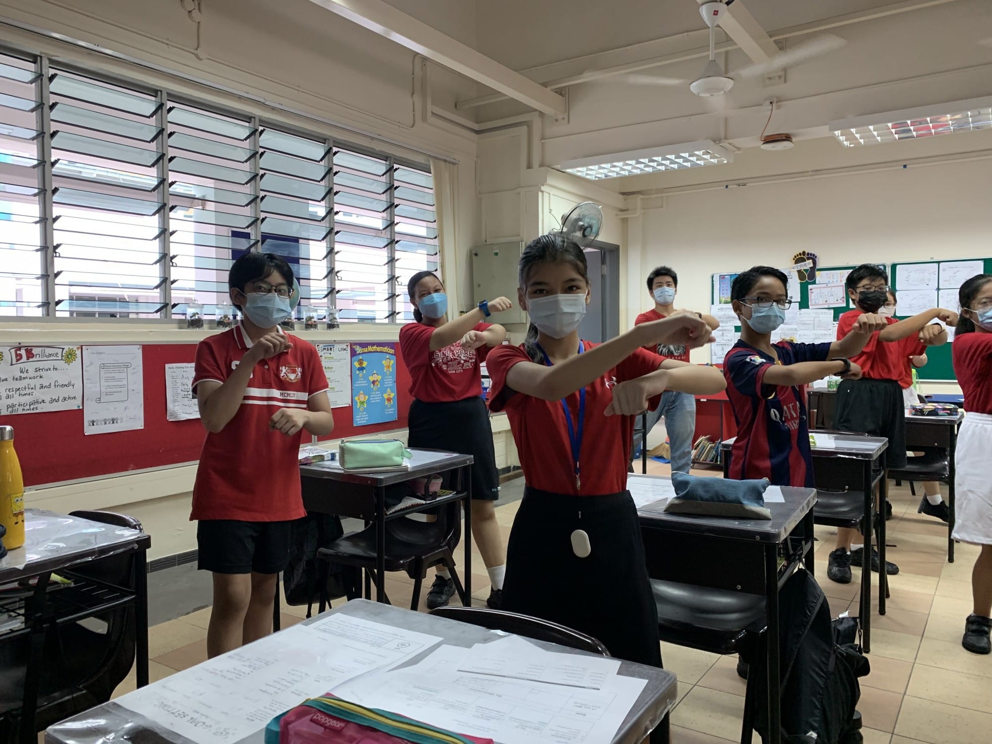 Students in red uniforms and face masks stretch their arms forward in a classroom with posters on the walls.
