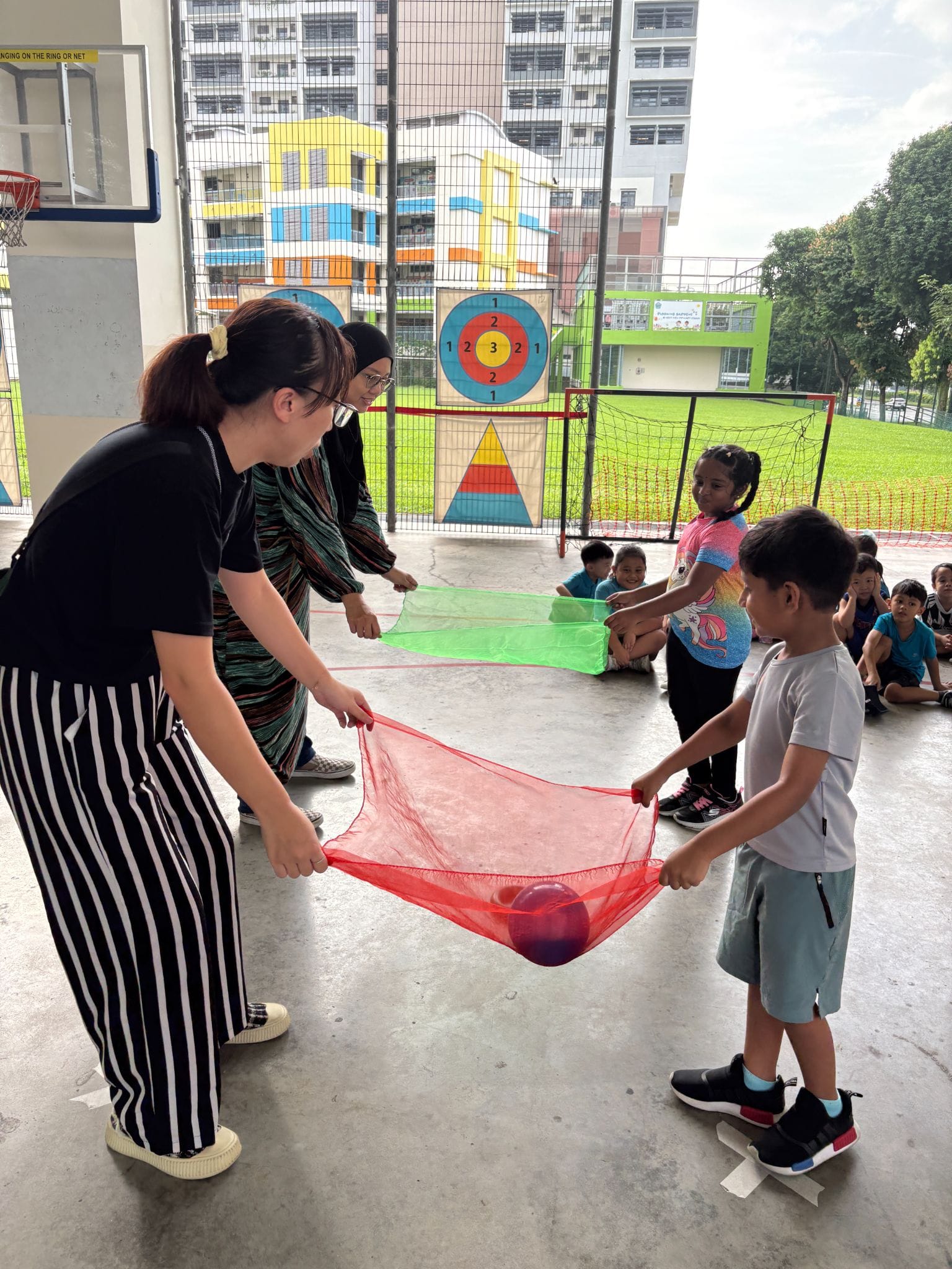 Adults and children hold red mesh fabric with a ball during an outdoor school activity, with other kids watching.