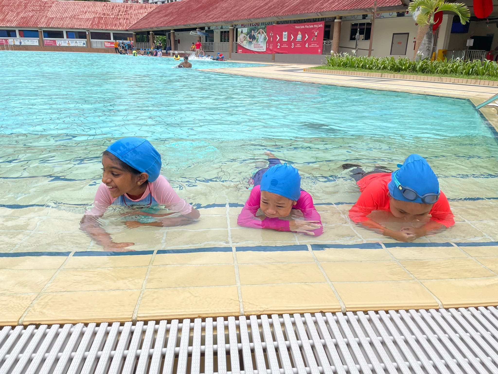 Three children wearing blue swim caps lie at the shallow edge of an outdoor public swimming pool.