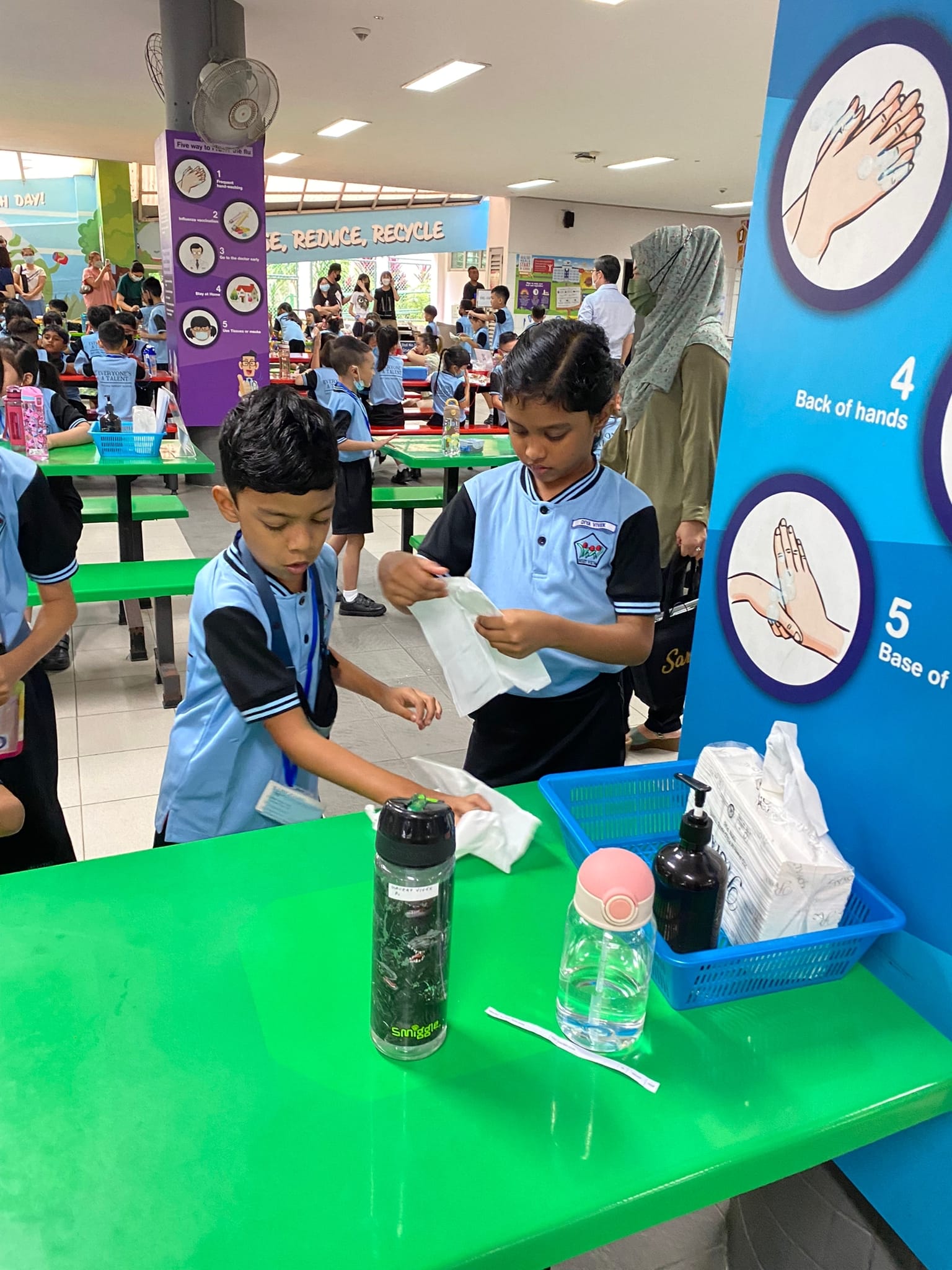 Two young students in blue uniforms handle tissue packets at a green table near a hand-washing steps poster in a school canteen.