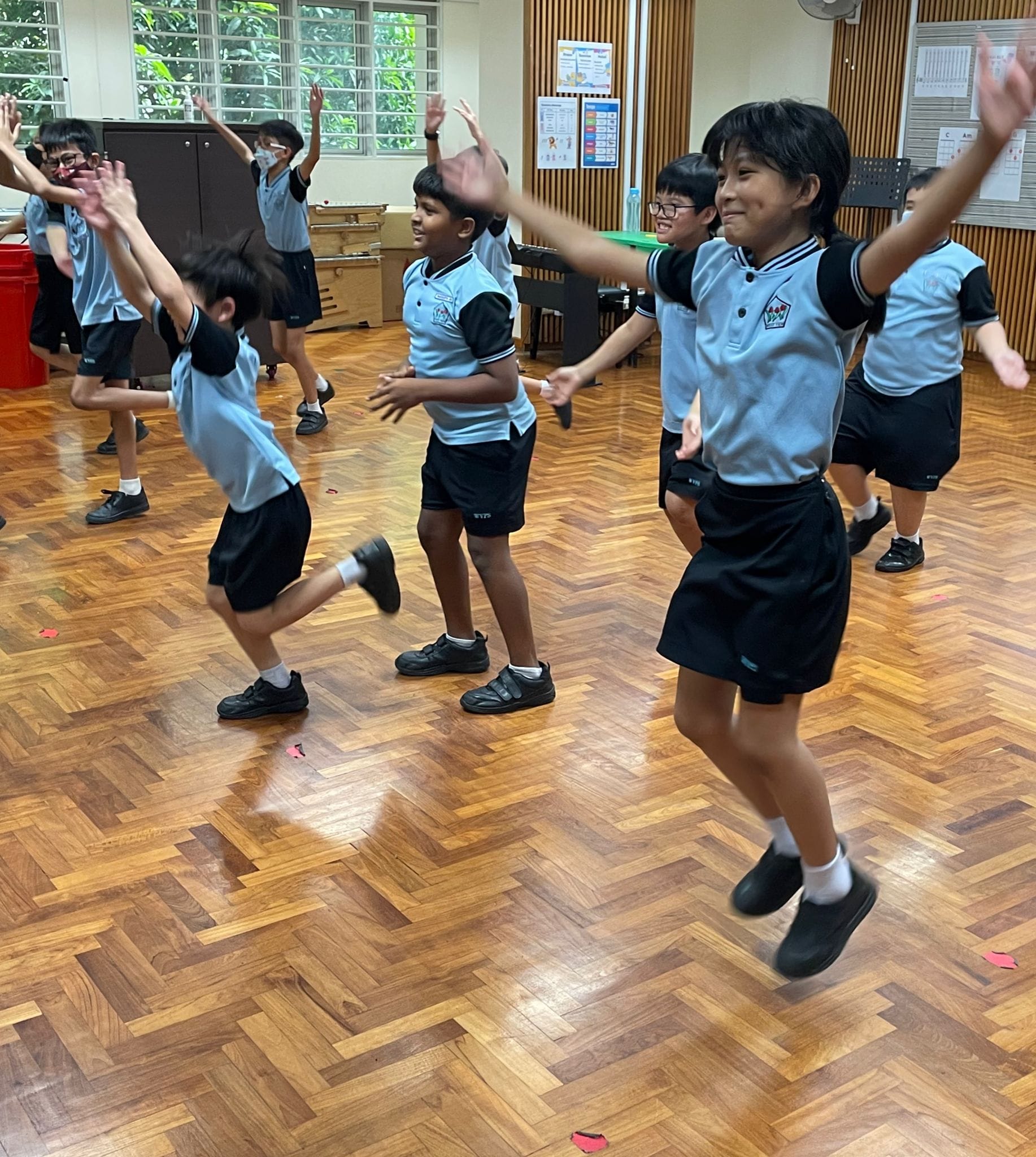 Several school children in blue and black uniforms jump and dance energetically on a wooden parquet floor indoors.