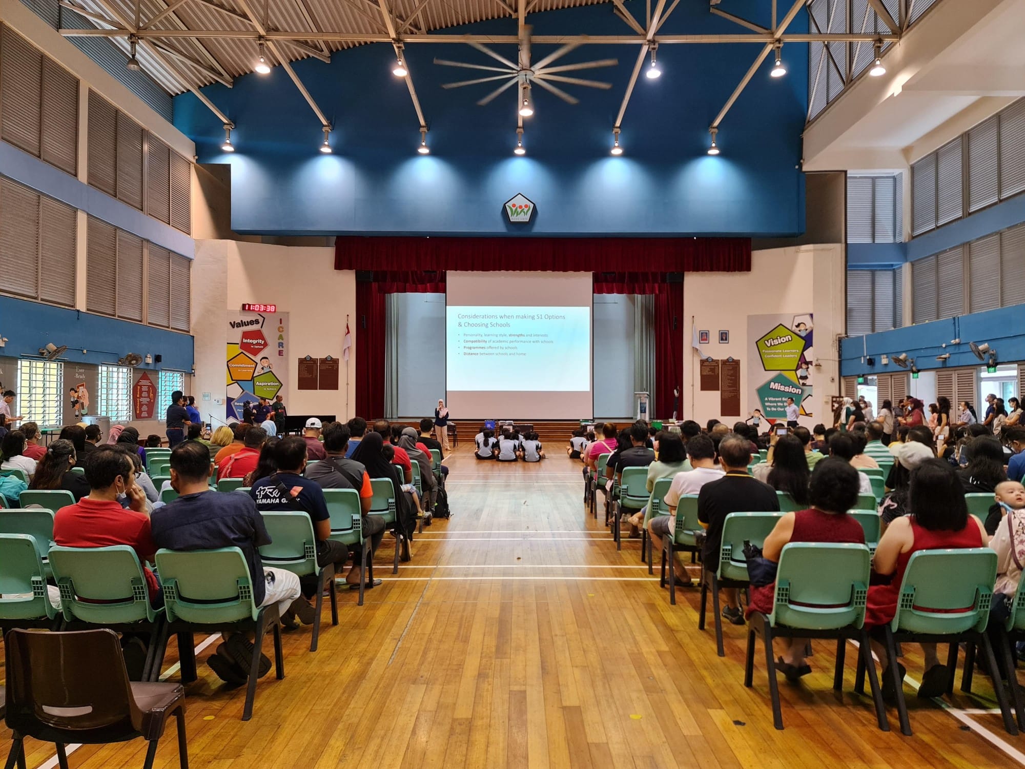 A school hall filled with seated attendees watching a presenter on stage with a projected slideshow about S1 options.