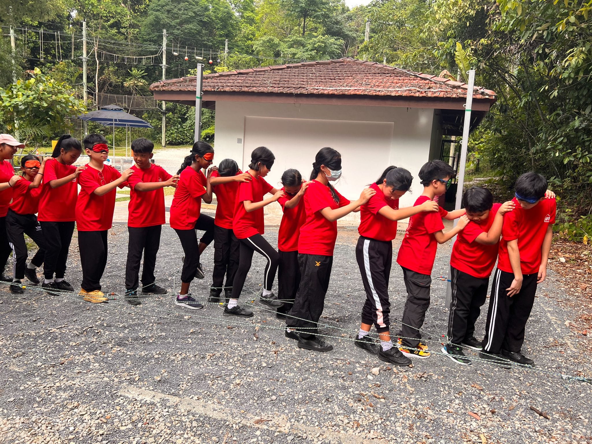 Children in red shirts with blindfolds hold each other's shoulders in a line, navigating a rope obstacle outdoors.