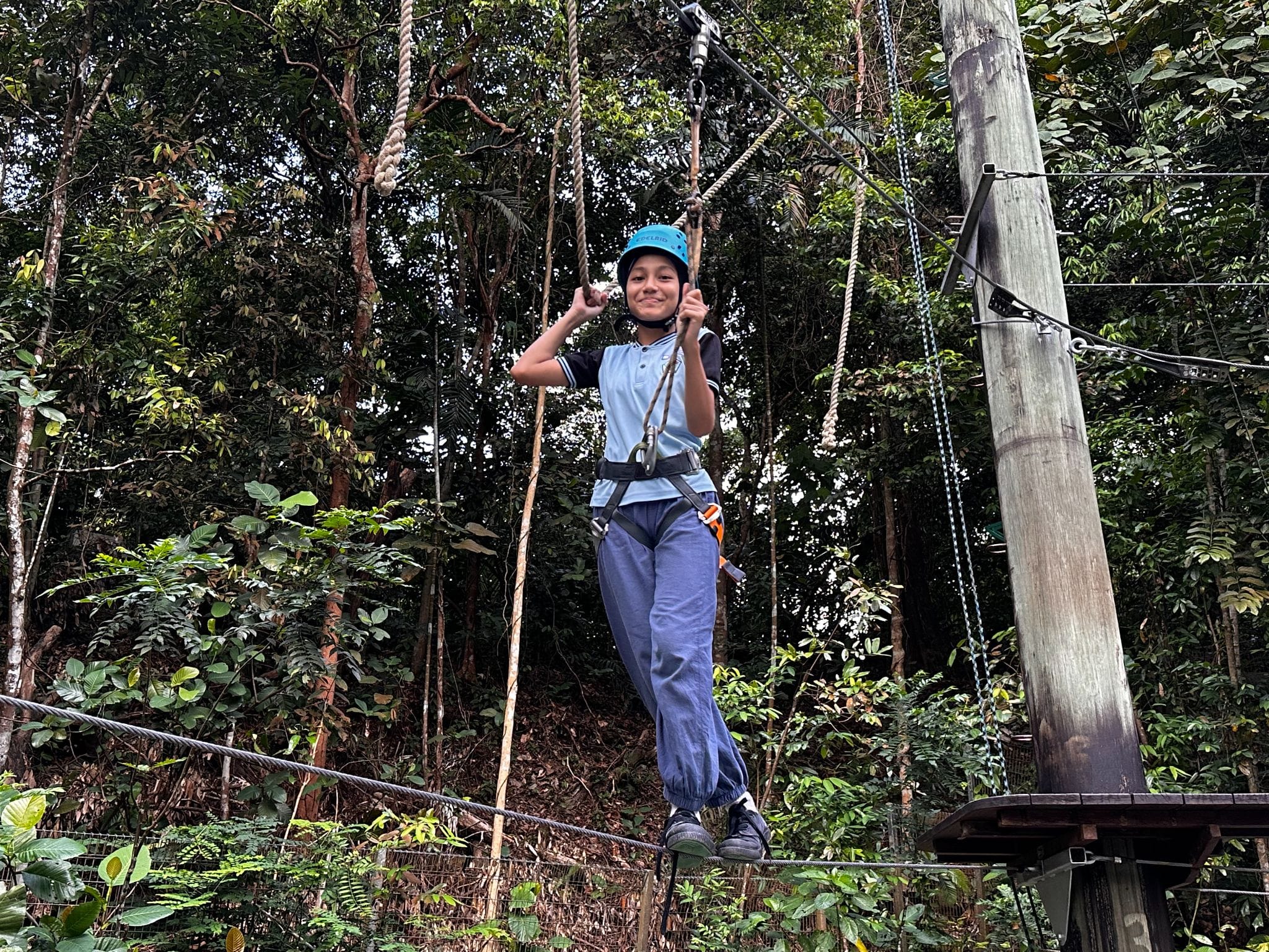 A smiling boy wearing a blue helmet and harness hangs on ropes at an outdoor treetop adventure course.