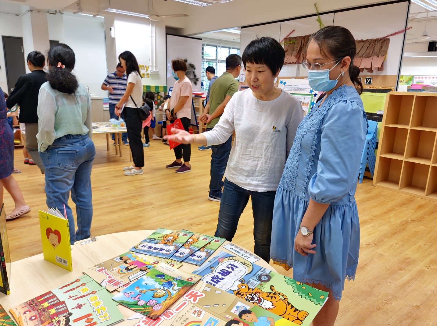 Two women look at colorful Chinese children's books on a table in a busy indoor classroom setting.