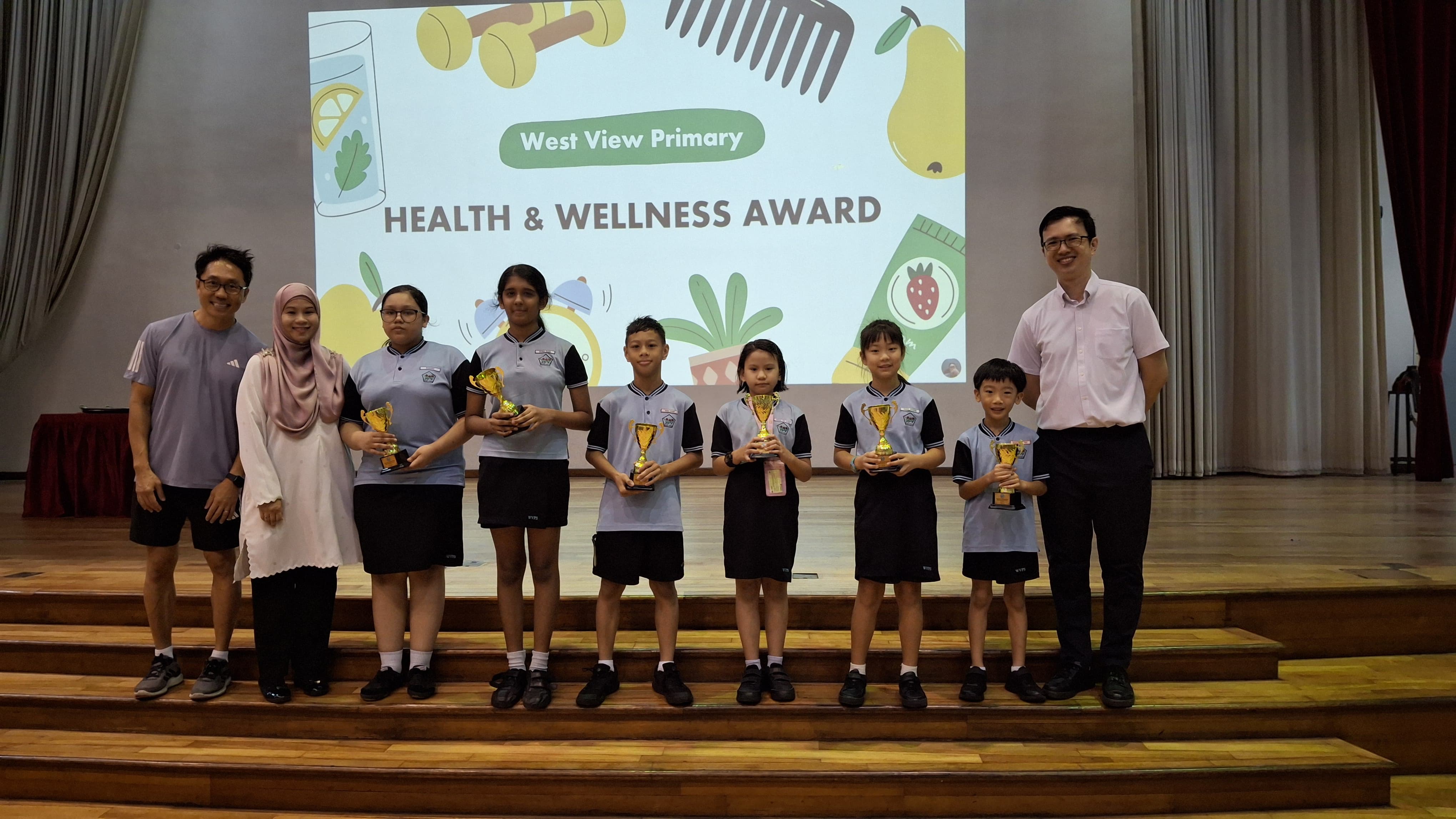 Students holding gold trophies on stage at West View Primary Health and Wellness Award ceremony with two adults.