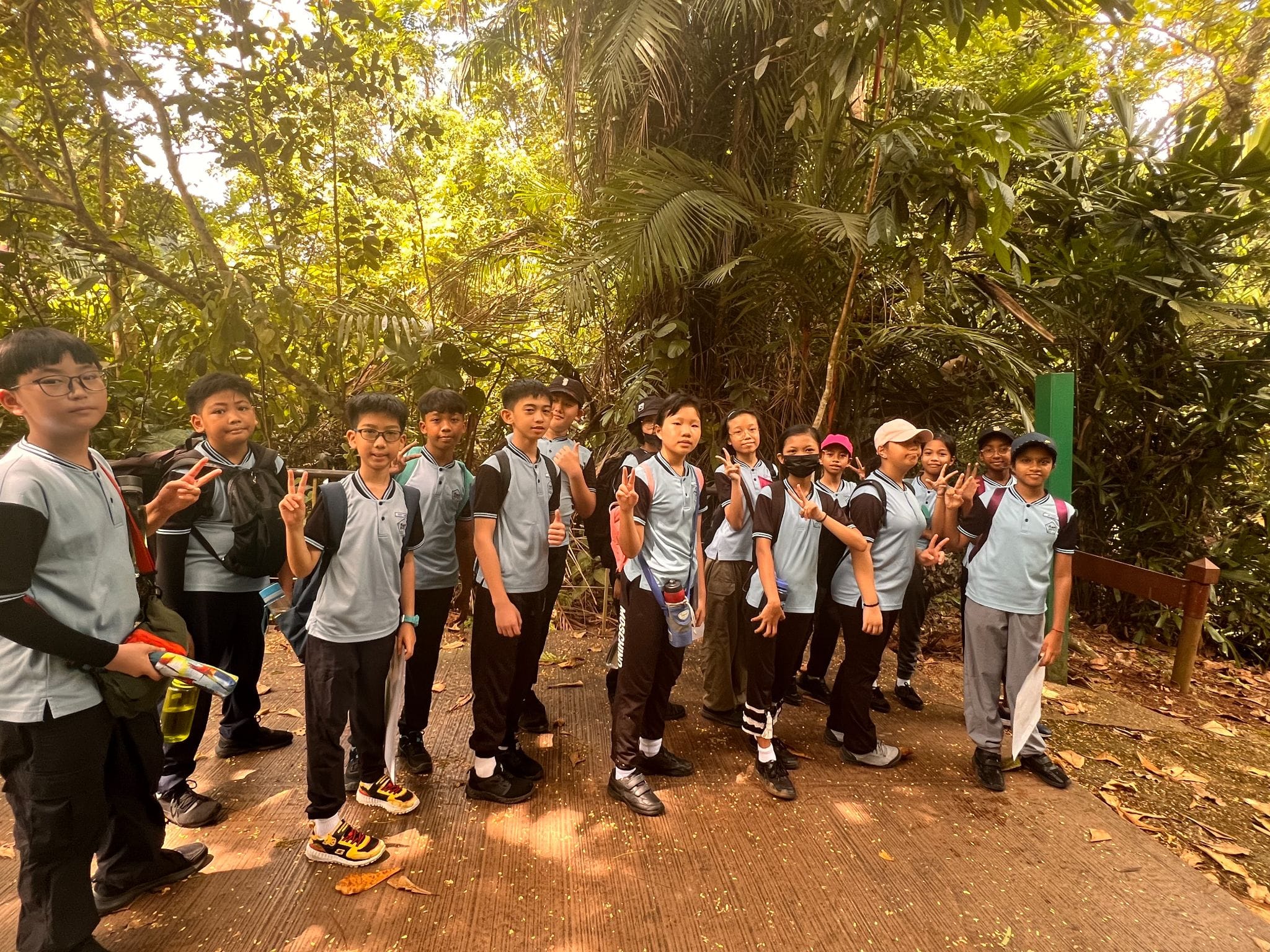 A group of school children in matching light blue uniforms pose with peace signs on a forest trail.