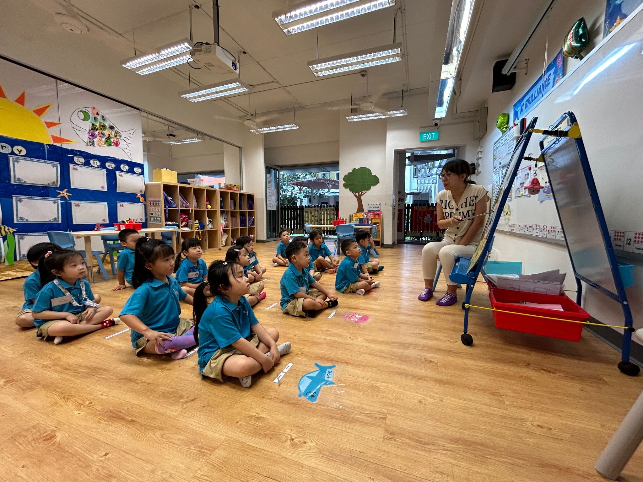 A teacher sits beside a whiteboard while young children in blue uniforms sit on a classroom floor listening attentively.