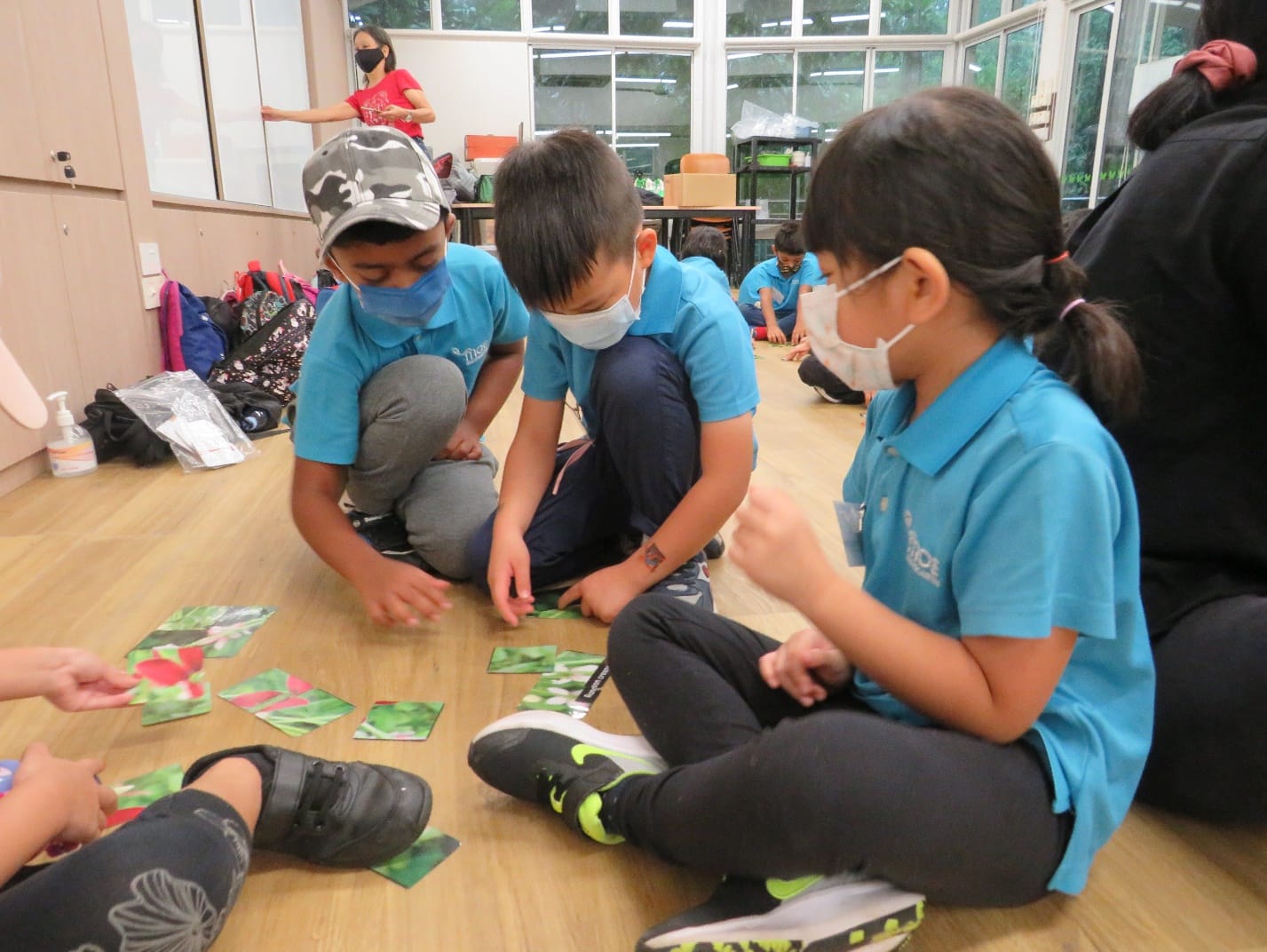 Children in blue uniforms and face masks sit on a wooden floor sorting picture cards in a classroom.