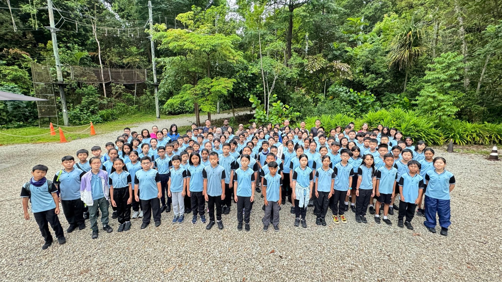 Large group of students in light blue and black uniforms posing outdoors on gravel surrounded by dense green trees.