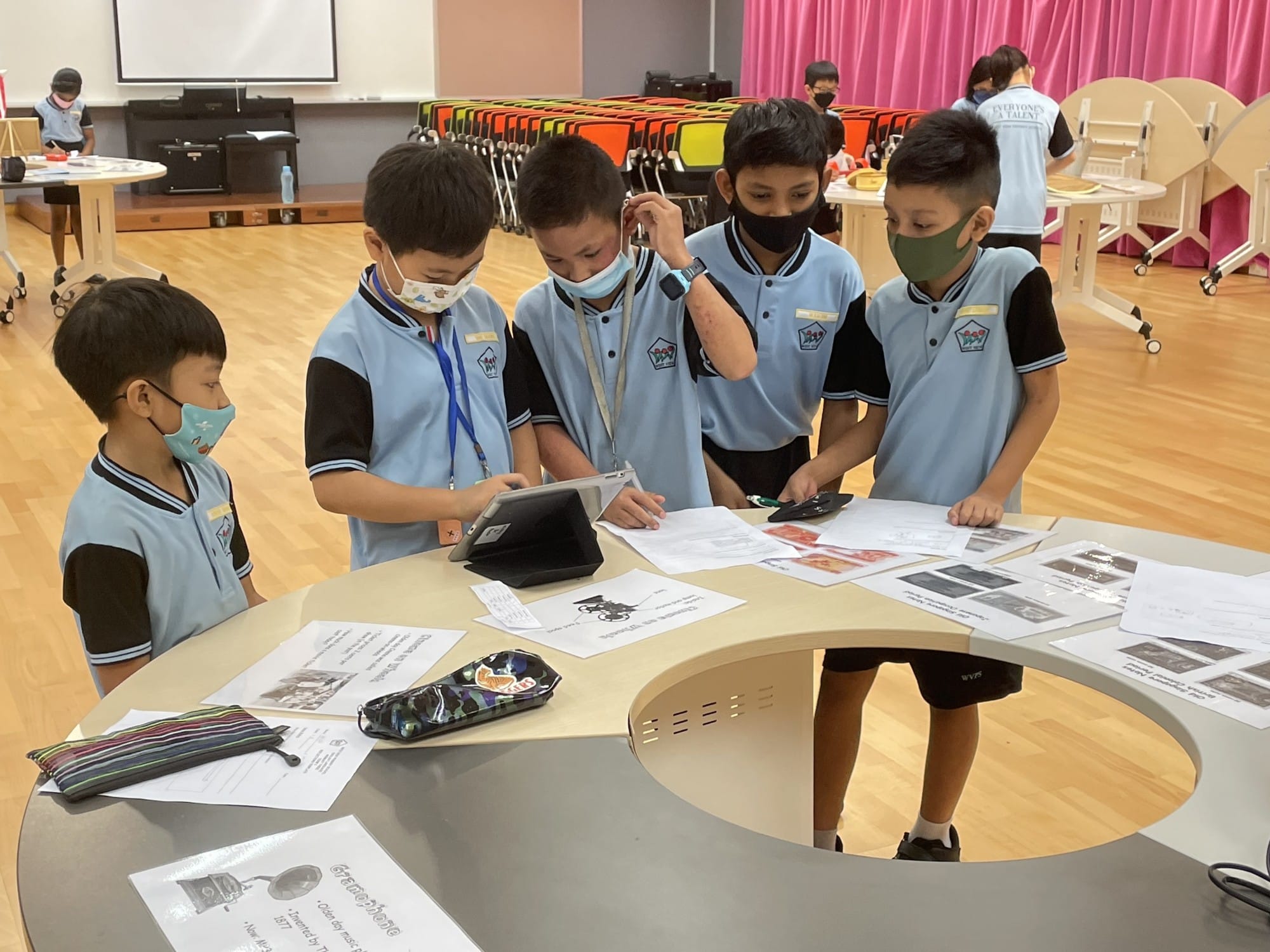 Four masked schoolboys in blue uniforms gather around a round table with papers and a tablet in a classroom.