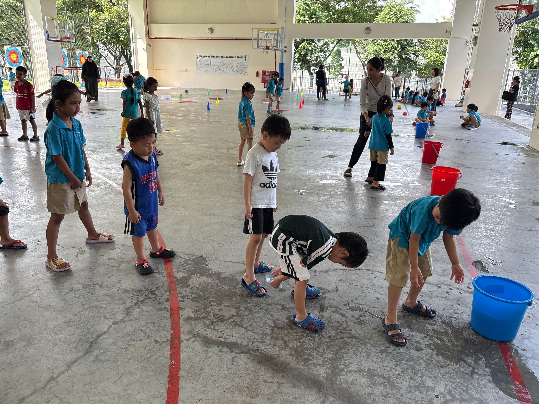 Young children in teal uniforms participate in an outdoor school activity with colored buckets and cones on a covered court.