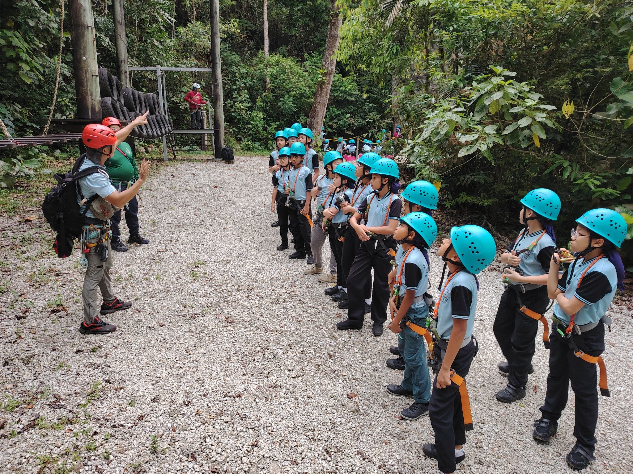 An instructor in a red helmet briefs a group of children in teal helmets and harnesses at an outdoor adventure course.