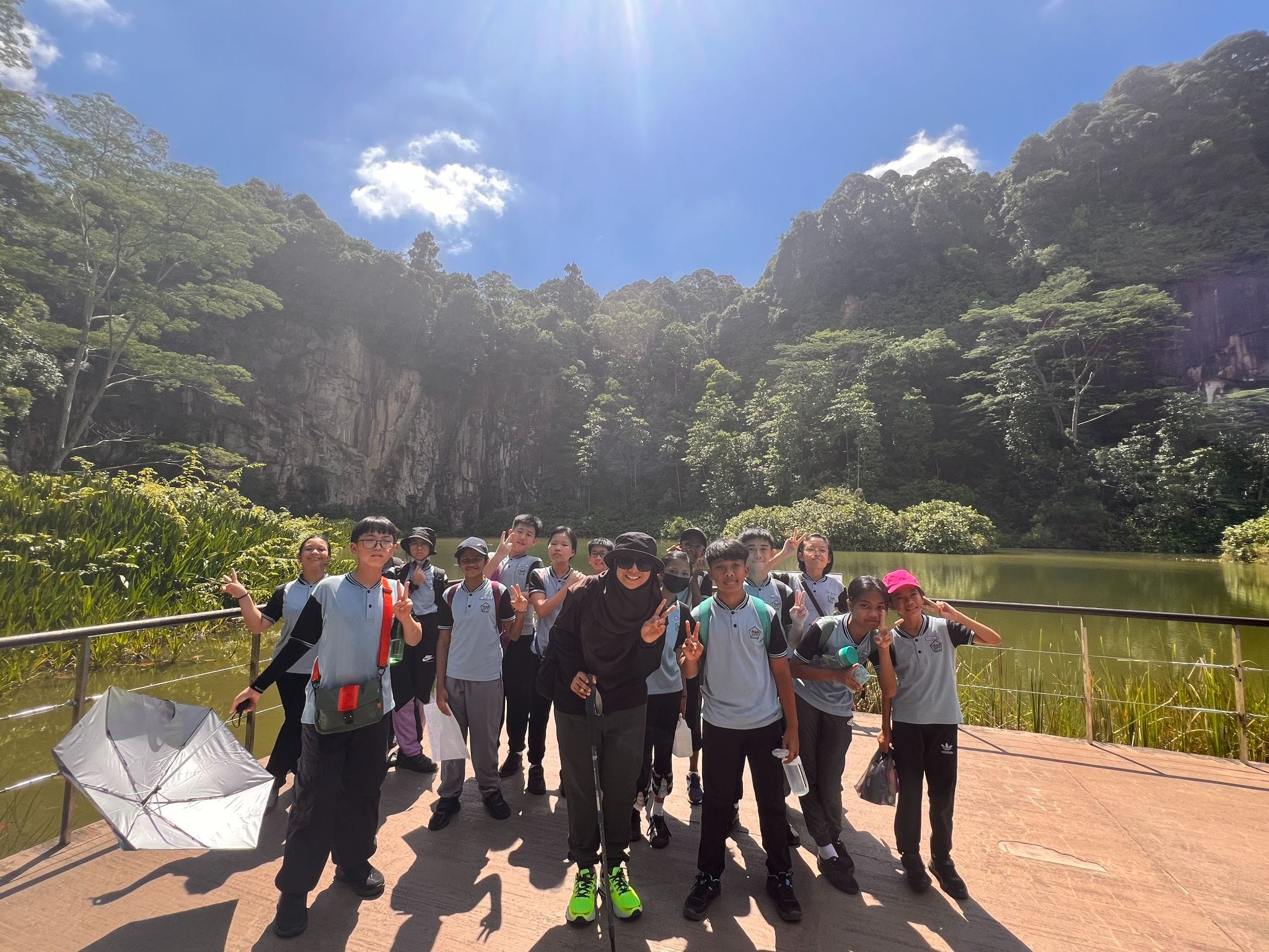 A group of students in matching uniforms pose on a platform by a green lake surrounded by forested limestone cliffs.