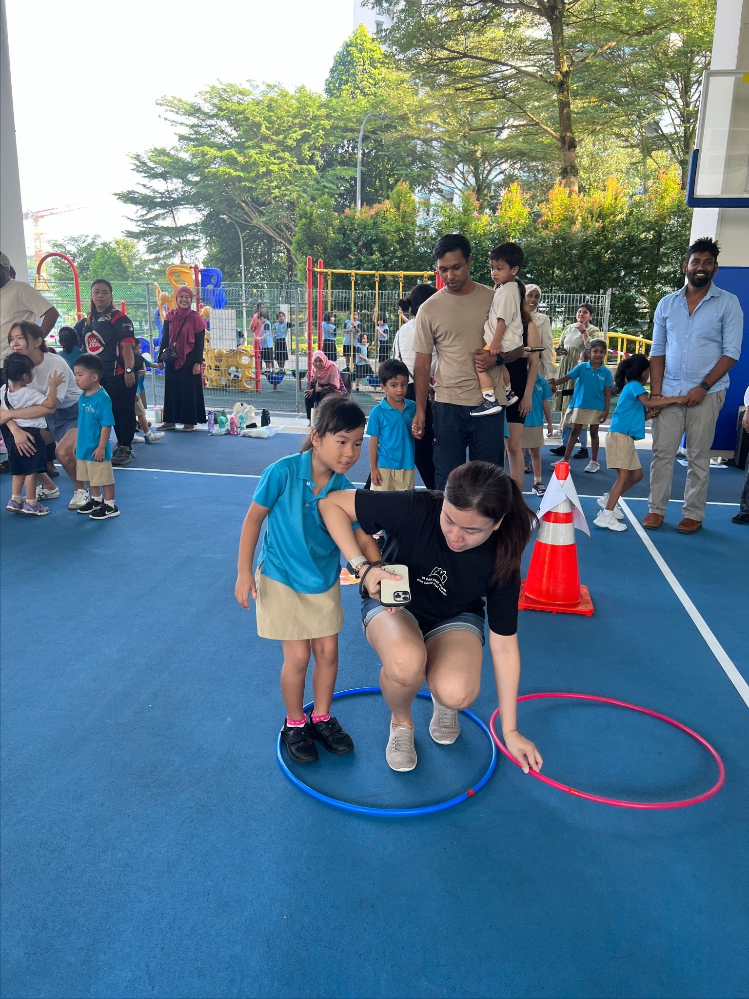 An adult crouches beside a young girl with hula hoops on a blue outdoor court surrounded by children and parents.