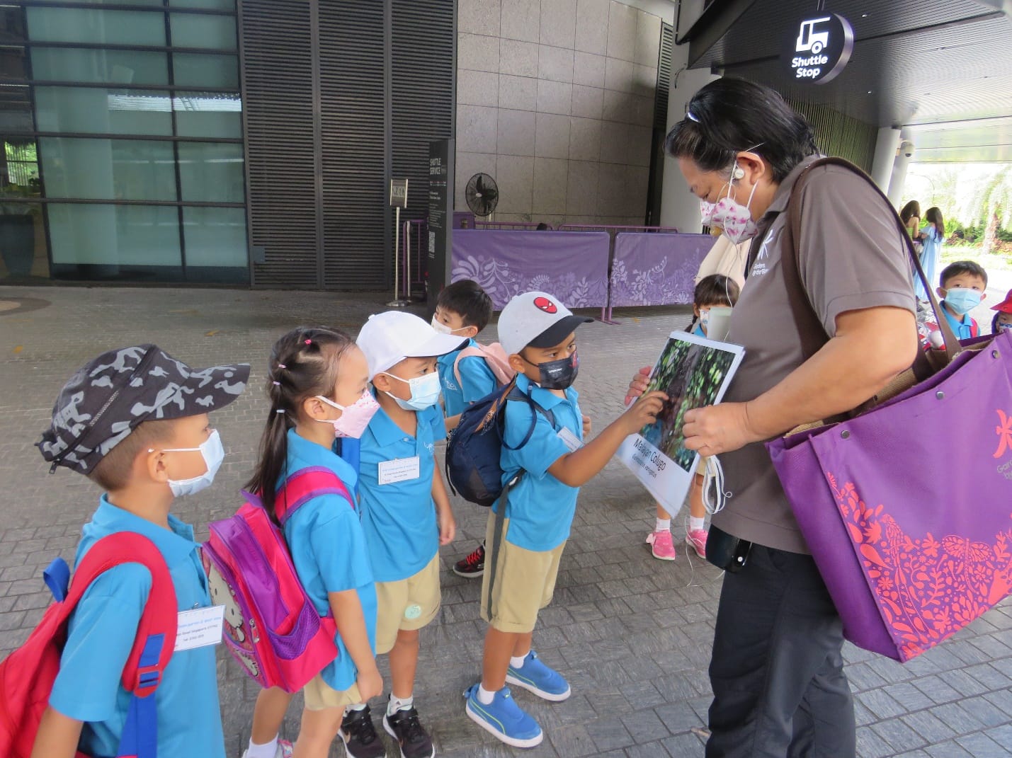 An adult guide shows a nature photo to young masked schoolchildren in blue uniforms outside a shuttle stop building.