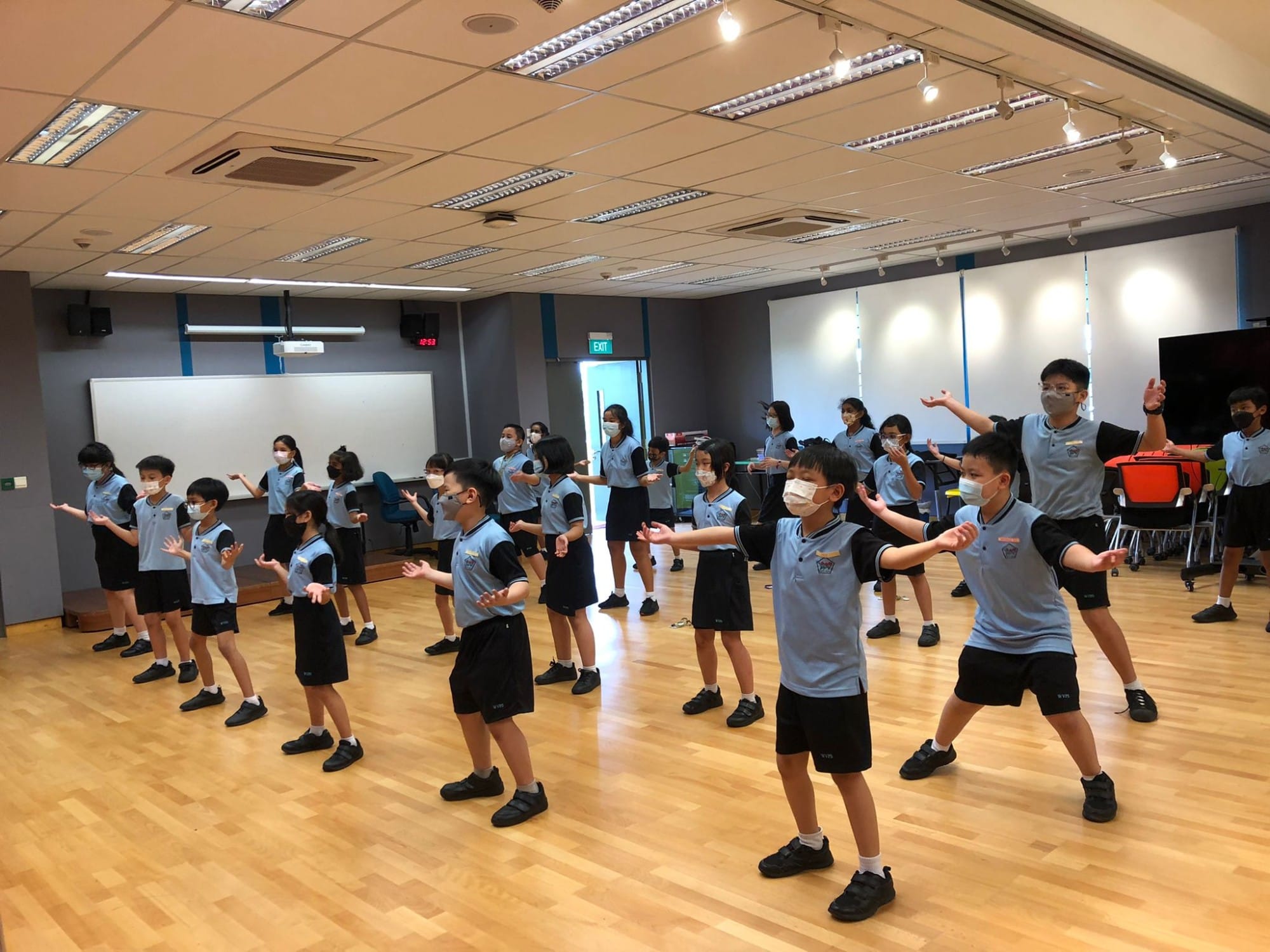 Students in blue uniforms and face masks practice a dance routine in a school activity room with wooden floors.