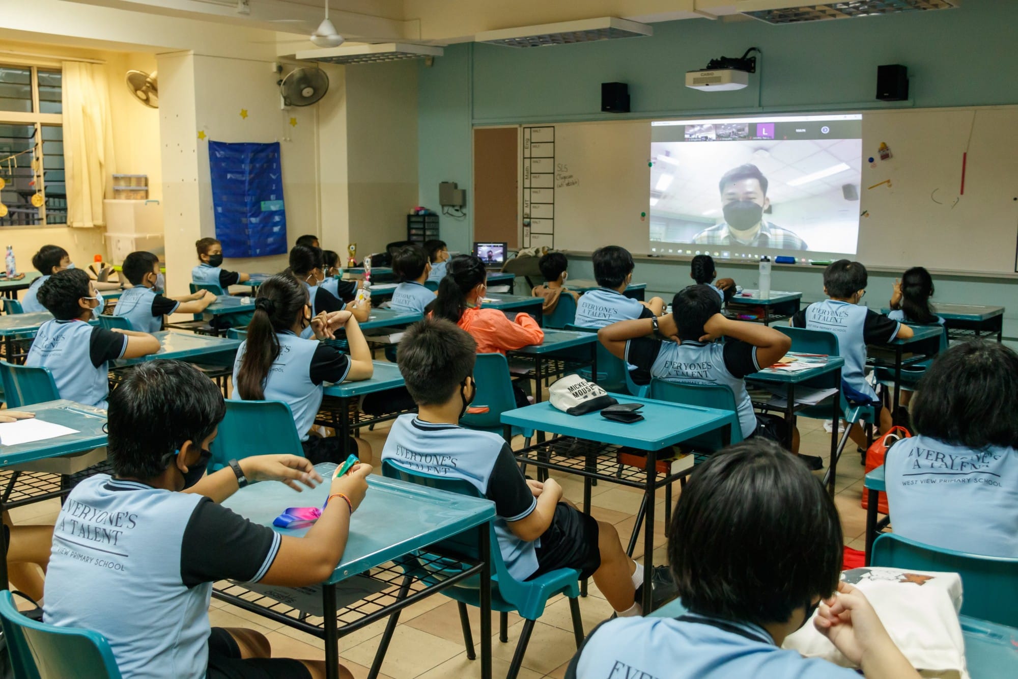 Students in blue uniforms sit at desks watching a masked person on a projected video call screen in a classroom.