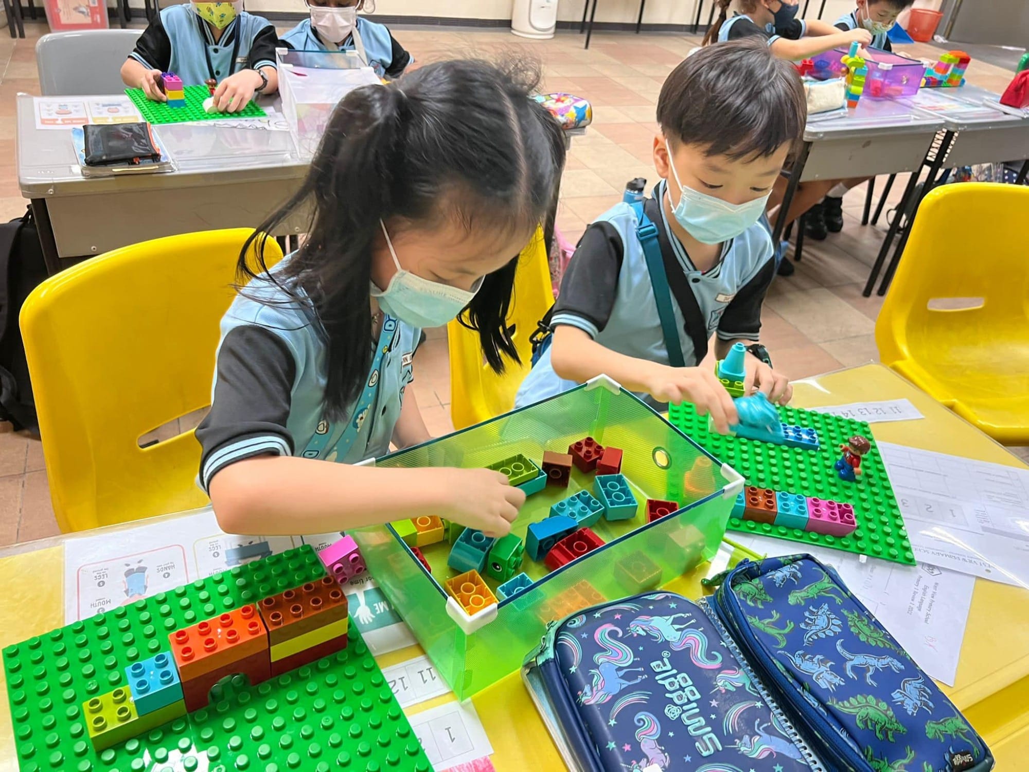 Two masked young children in school uniforms build with colorful Duplo blocks on green baseplates at a yellow table.