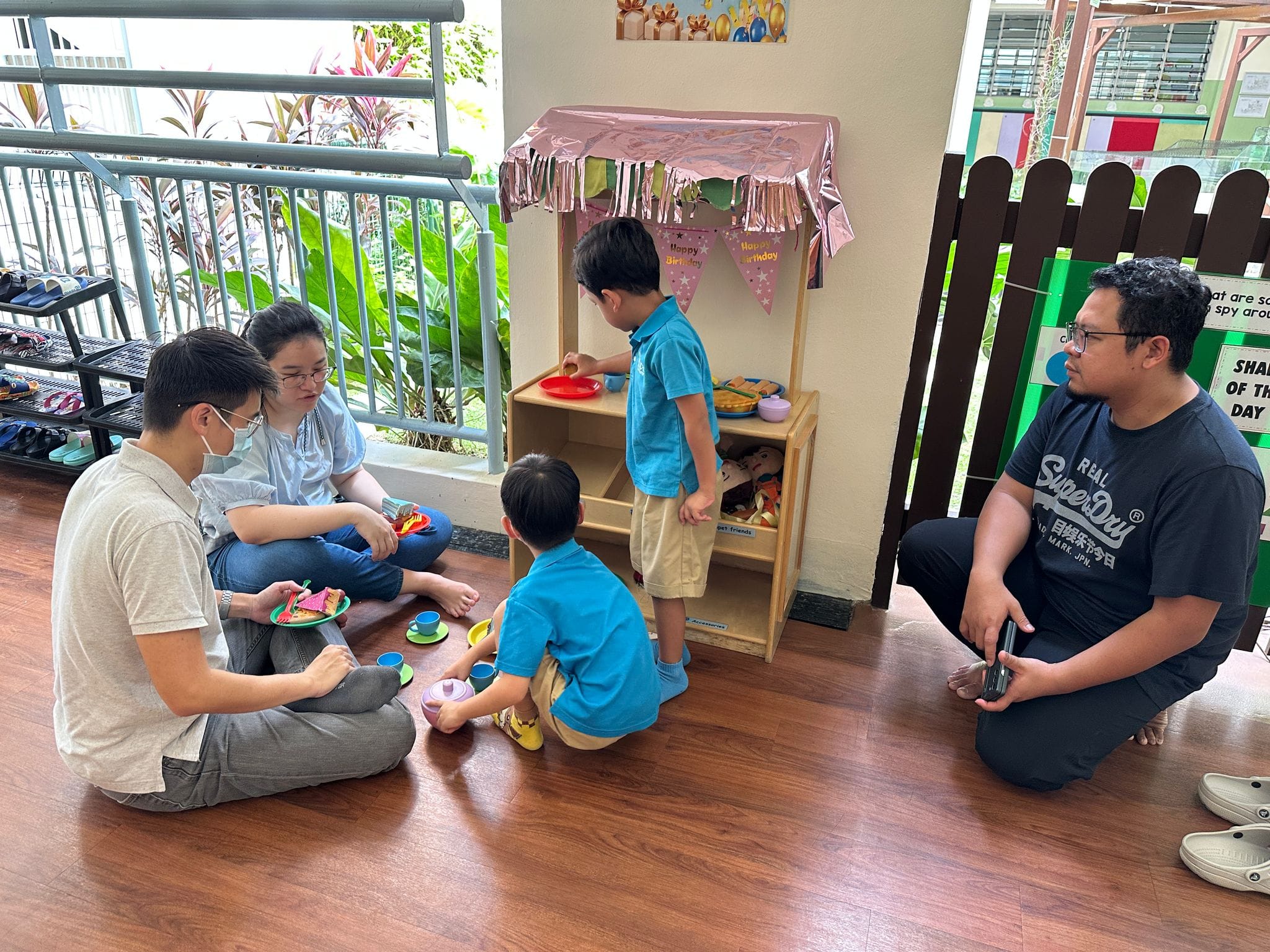 Two adults and two young children sit on a floor playing with toy food near a decorated wooden play kitchen.