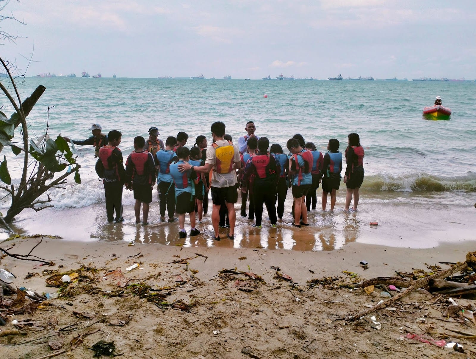 A group of people wearing life jackets stand together at the shoreline of a littered beach with ships visible at sea.