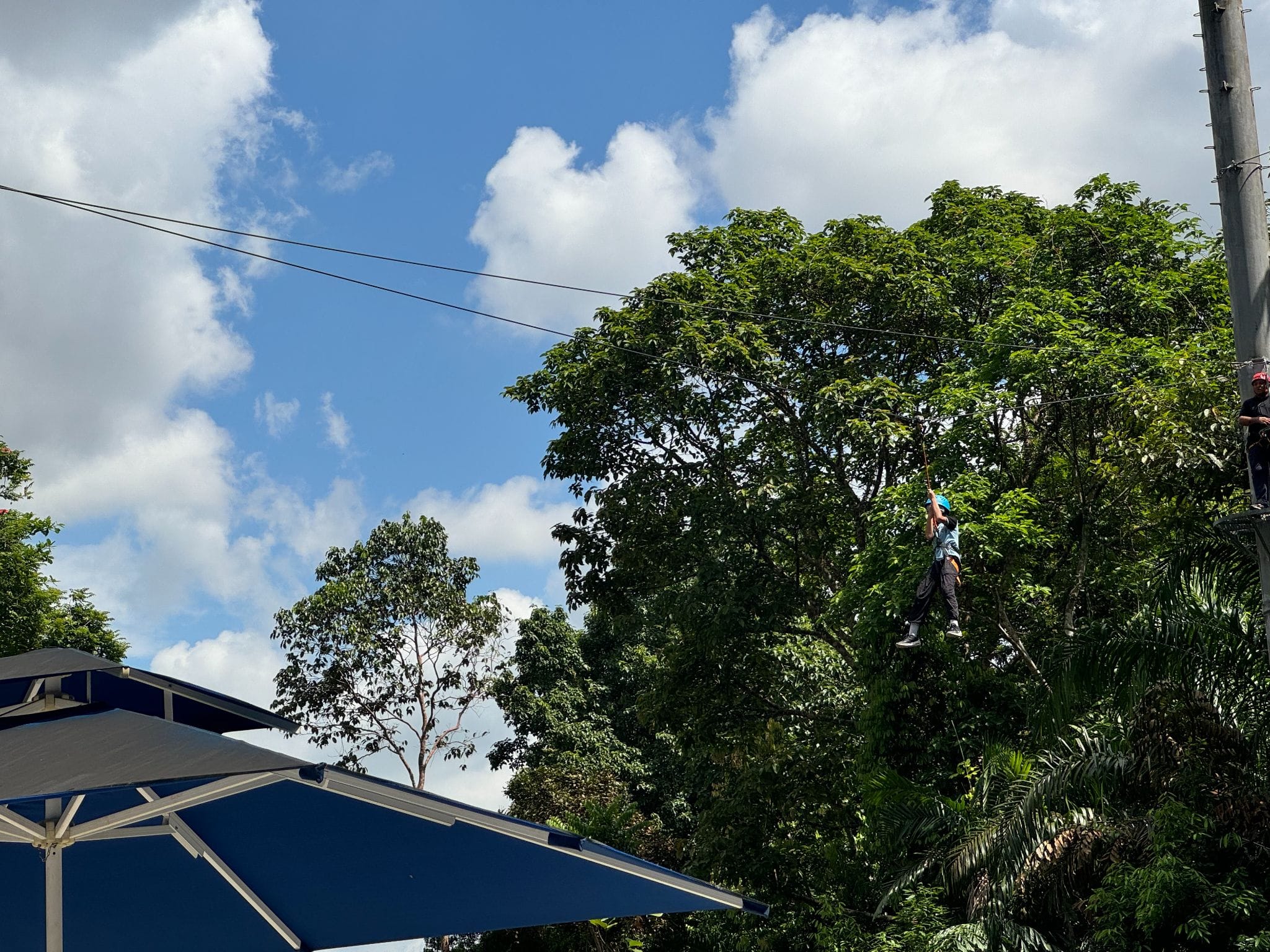 Person wearing a blue helmet zip-lining high above green trees under a partly cloudy blue sky.