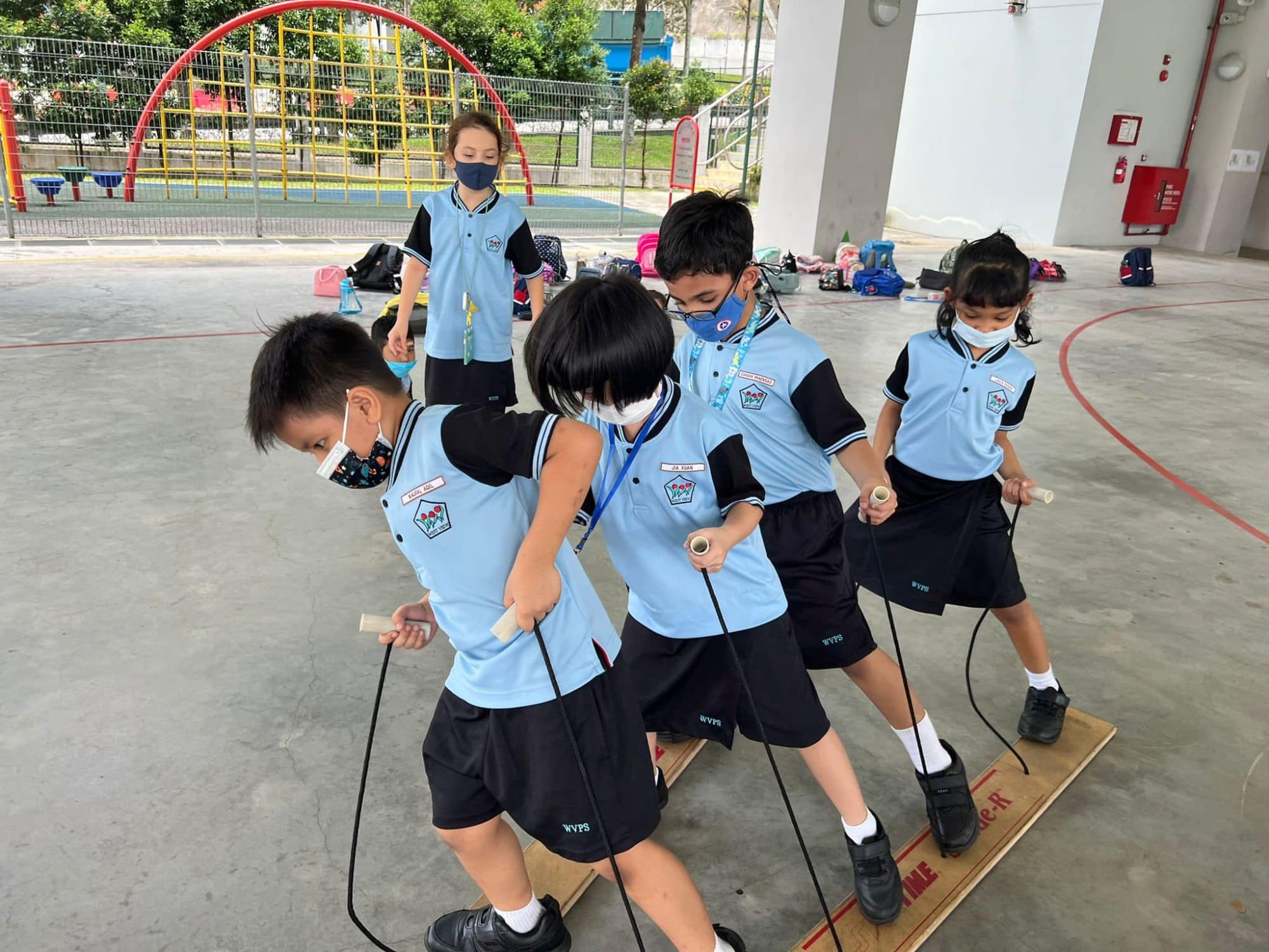 Four masked schoolchildren in blue uniforms balance together on a wooden board while holding rope handles outdoors.
