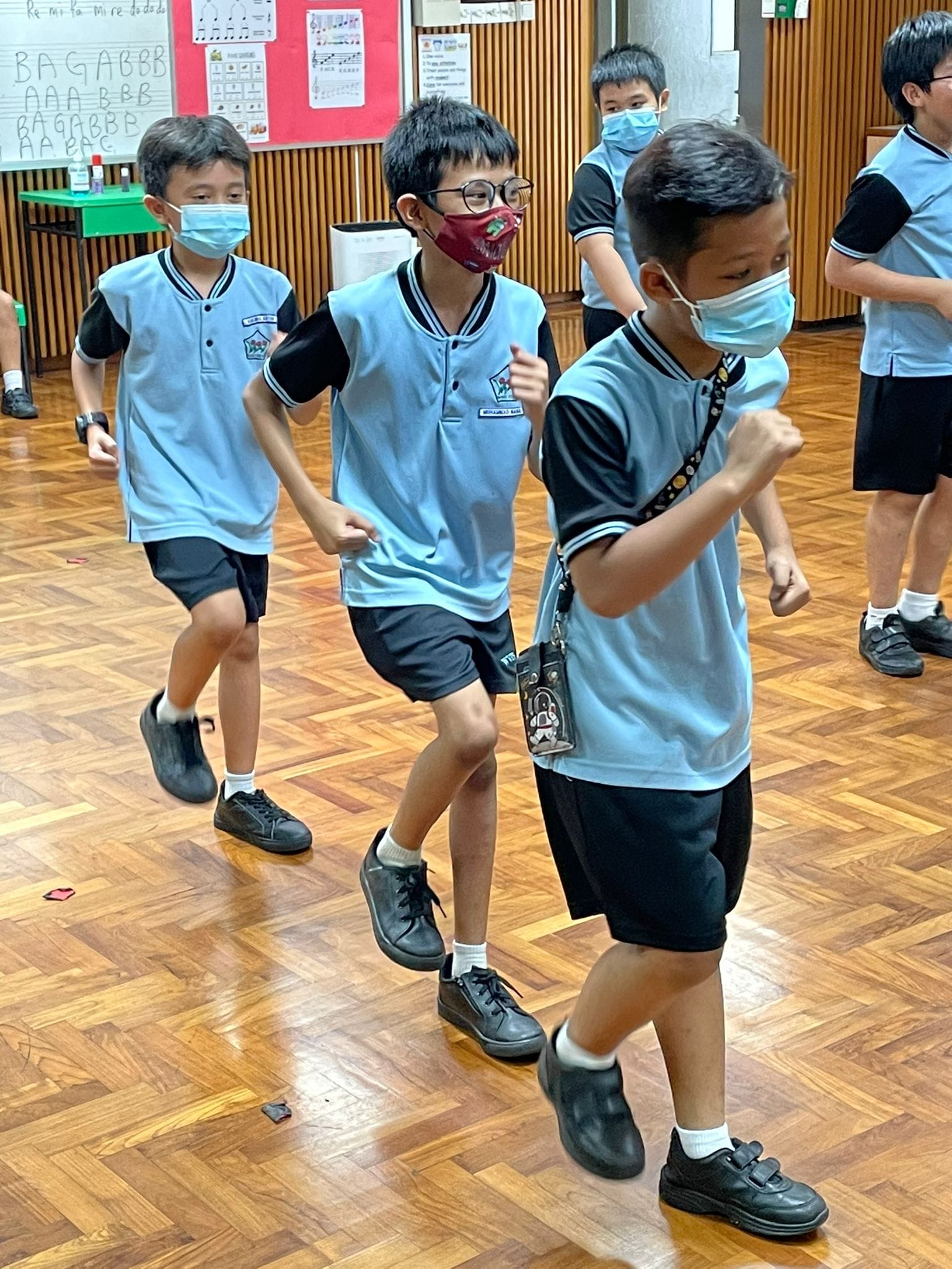 Several young students in blue school uniforms and face masks move actively on a wooden floor indoors.
