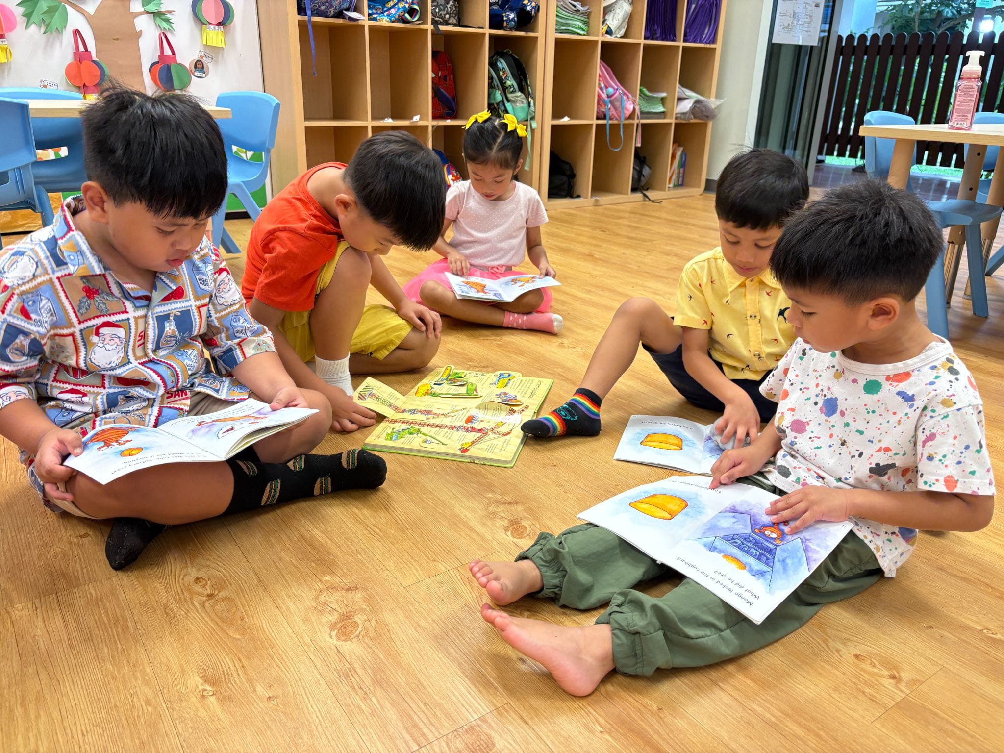 Five young children sit on a classroom floor reading colorful picture books together.