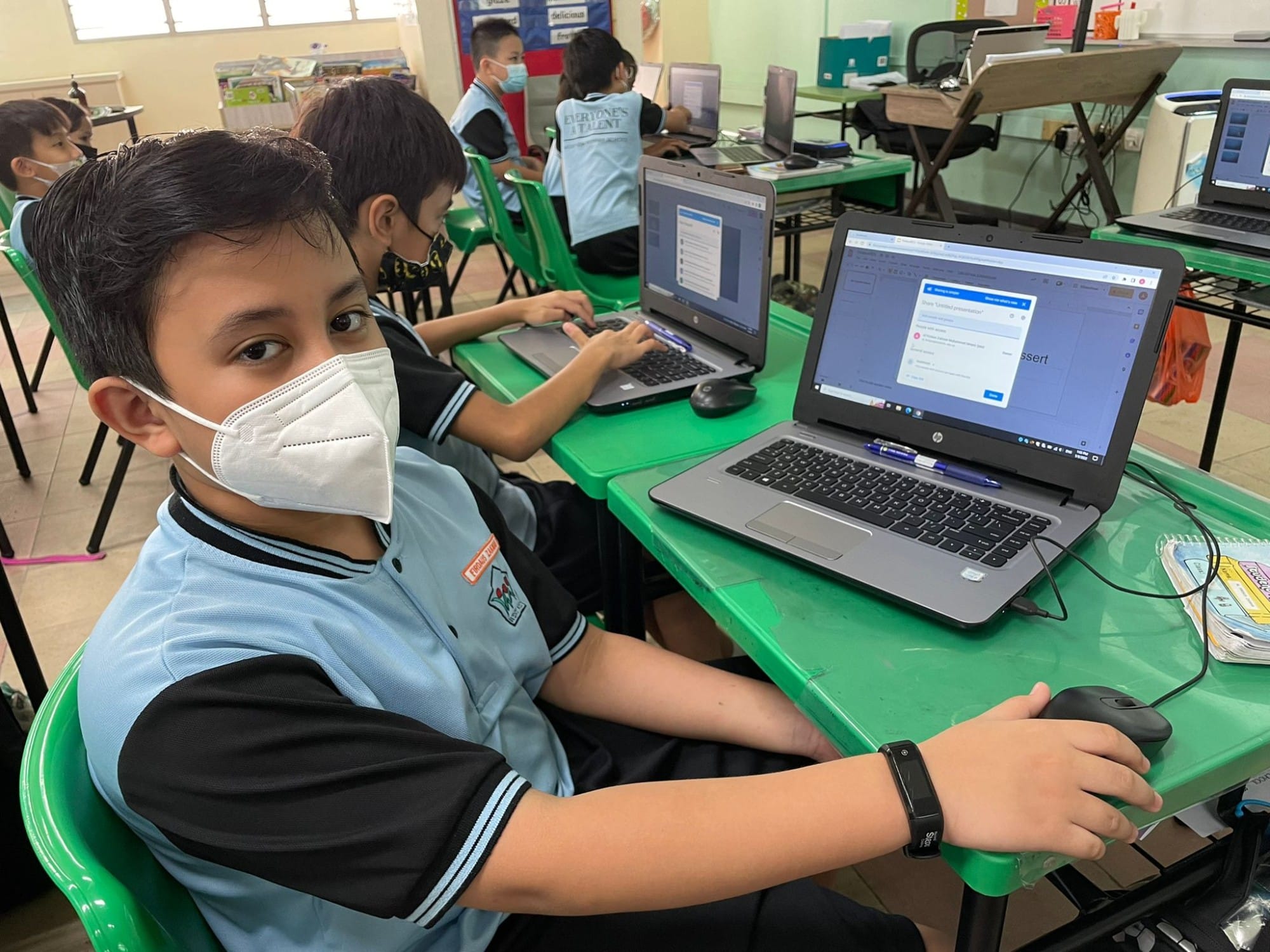 A masked boy in a blue school uniform sits at a green desk using a laptop in a classroom full of students.