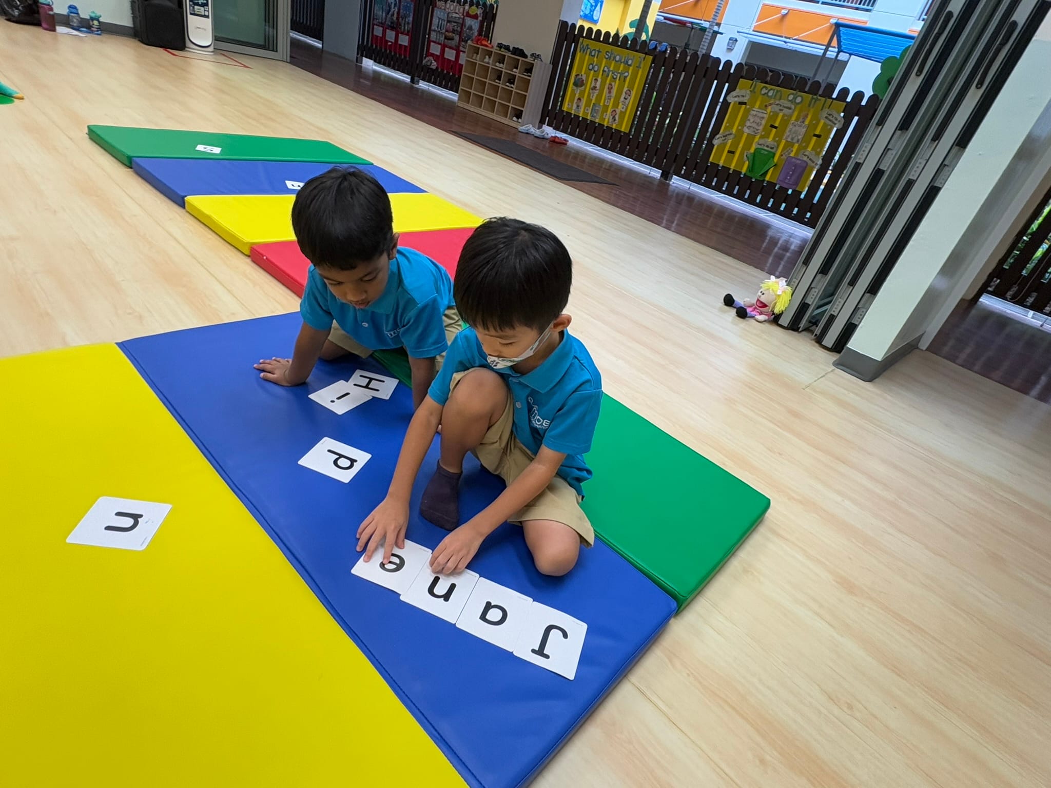 Two young boys in blue uniforms arrange letter cards spelling "Janep" on a colorful foam mat in a classroom.