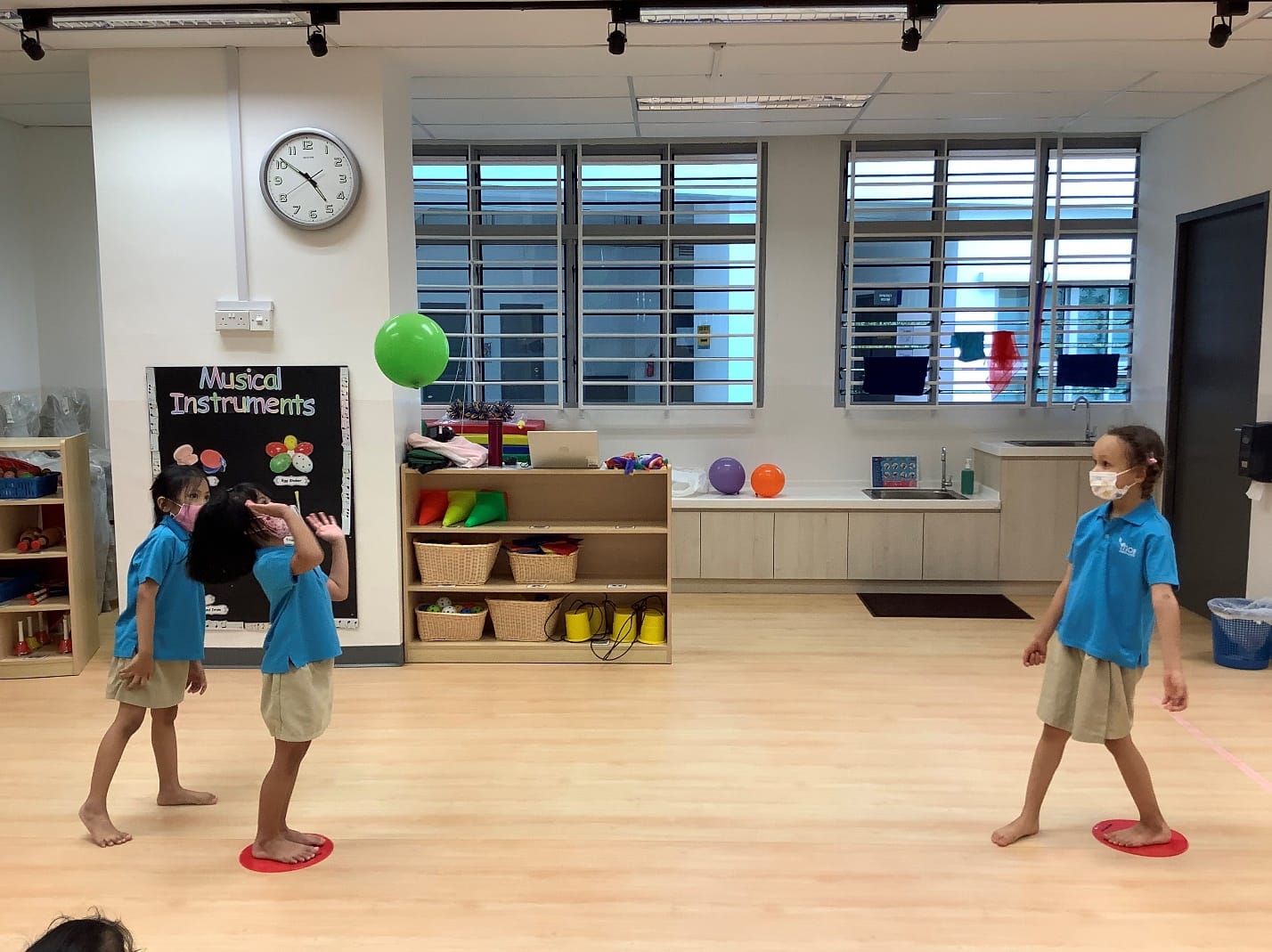 Three children in blue uniforms and masks play with a green balloon in a classroom with a Musical Instruments board.