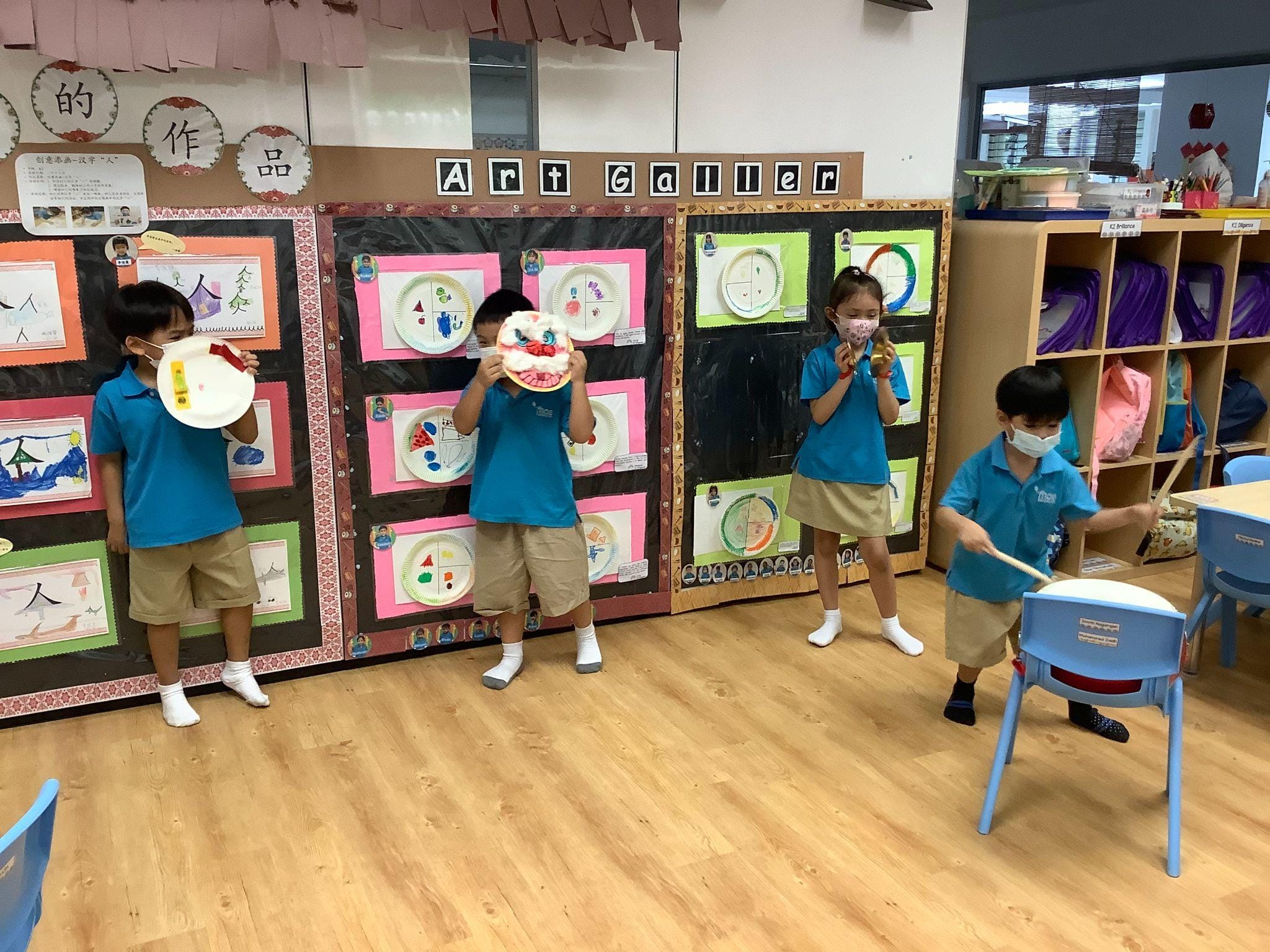 Four young students in blue uniforms hold decorated paper plates and drum in a classroom with an Art Gallery display.