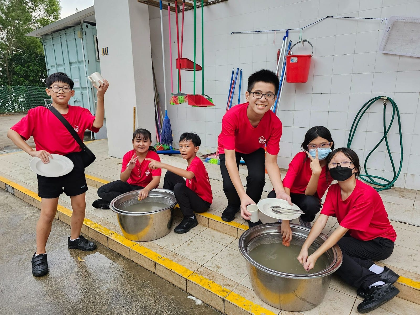 Six children in red shirts wash dishes in large metal basins outdoors, smiling at the camera.